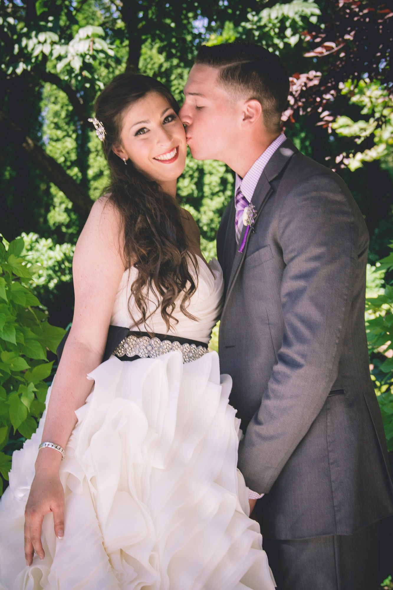 Groom in gray suit kisses smiling bride on cheek outdoors surrounded by lush green foliage.