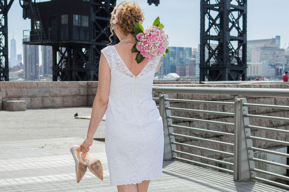 Bride in white lace dress carries pink hydrangea bouquet walking on urban waterfront pier.