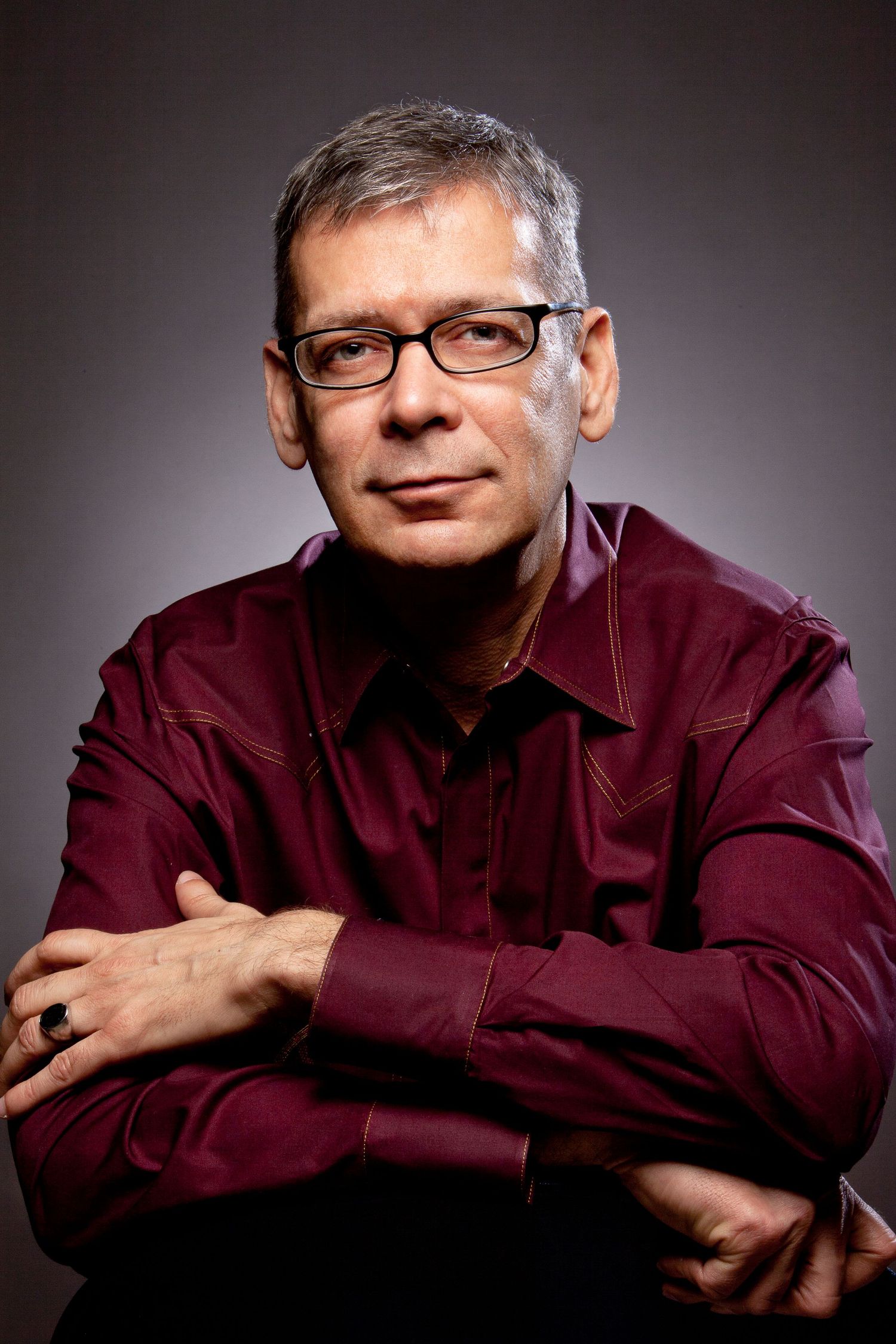 Middle-aged man with gray hair and glasses, arms crossed, wearing a dark red shirt, studio portrait.