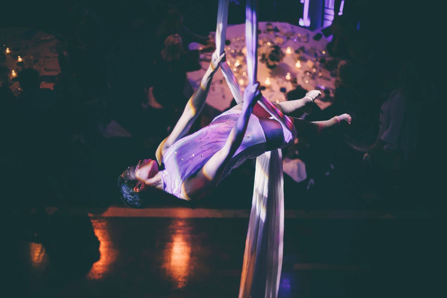 An aerial silk acrobat performs an inverted backbend pose in flowing white fabric at an event.