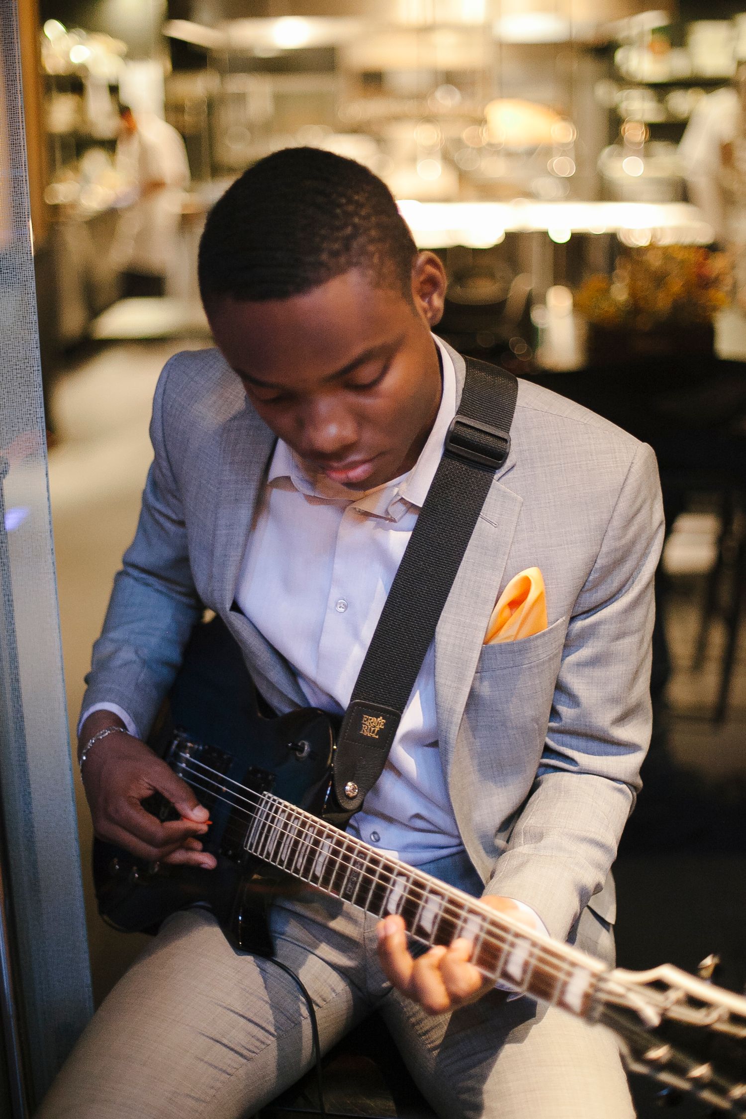 Young man in gray blazer playing black electric guitar at indoor venue.