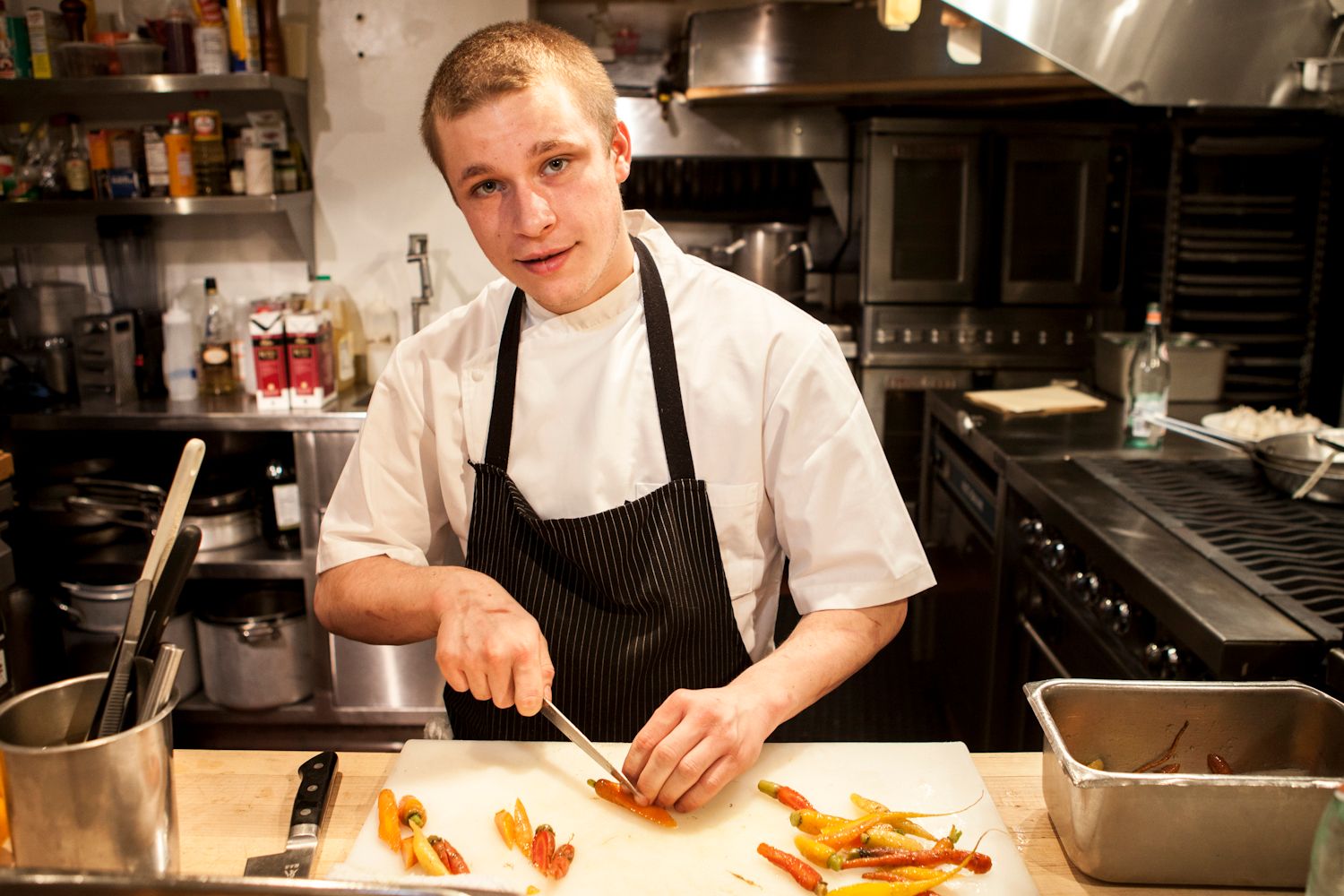 Young chef in white coat and black apron preparing colorful vegetables in a professional kitchen.