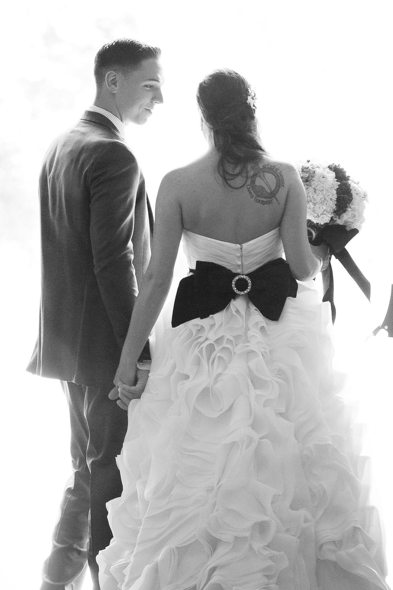 Black and white photo of bride in ruffled gown with black bow holding hands with smiling groom.