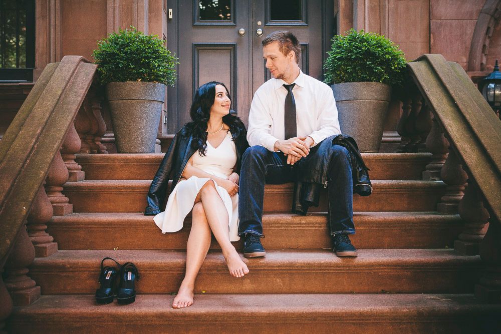 Couple sitting on brownstone steps gazing at each other, woman in white dress, man in white shirt and tie.