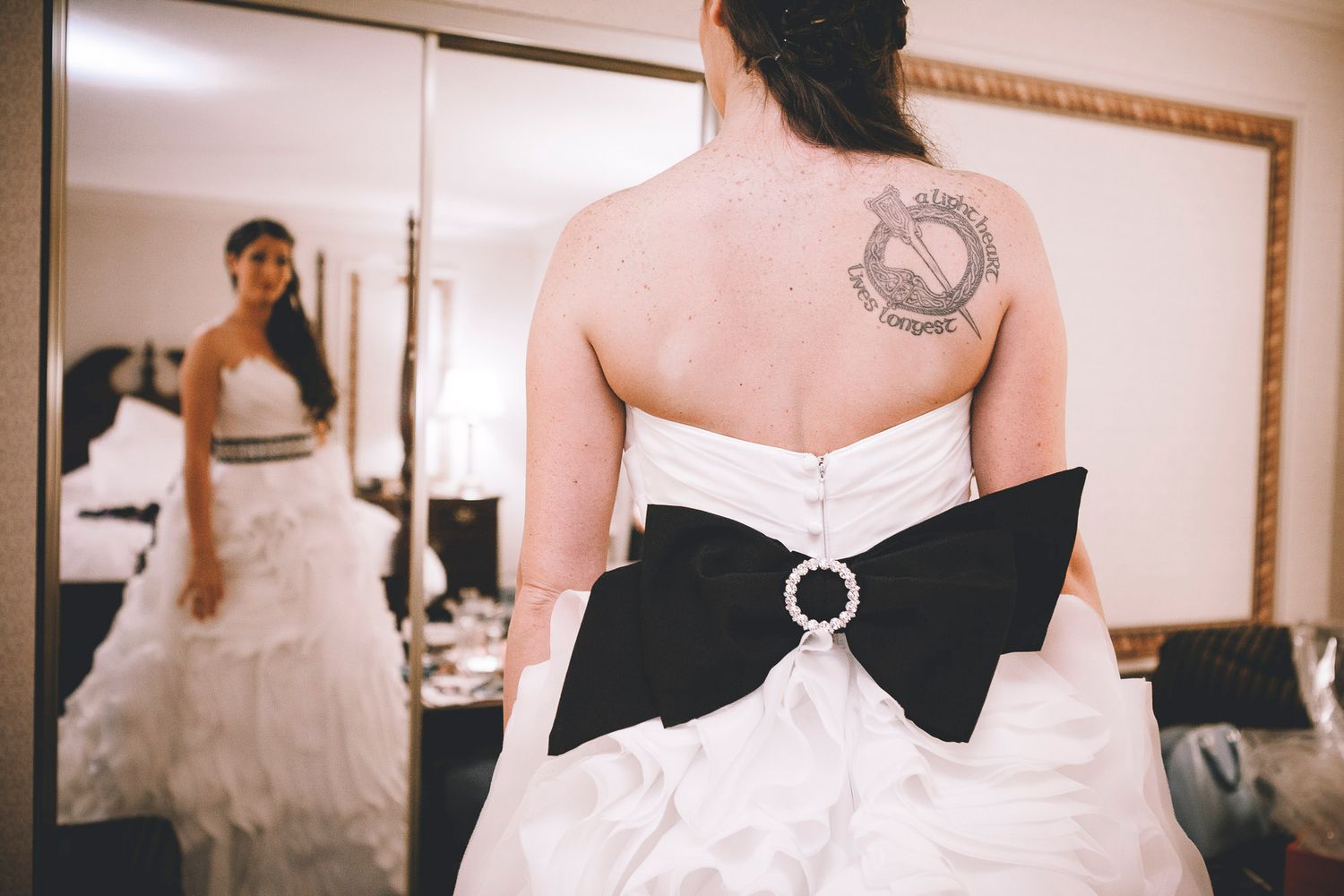 Bride in strapless white wedding gown with large black bow and circular brooch, back tattoo visible in mirror.