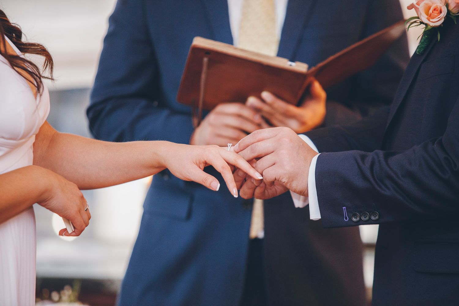 Groom placing wedding ring on bride's finger during ring exchange ceremony officiated by minister holding book.