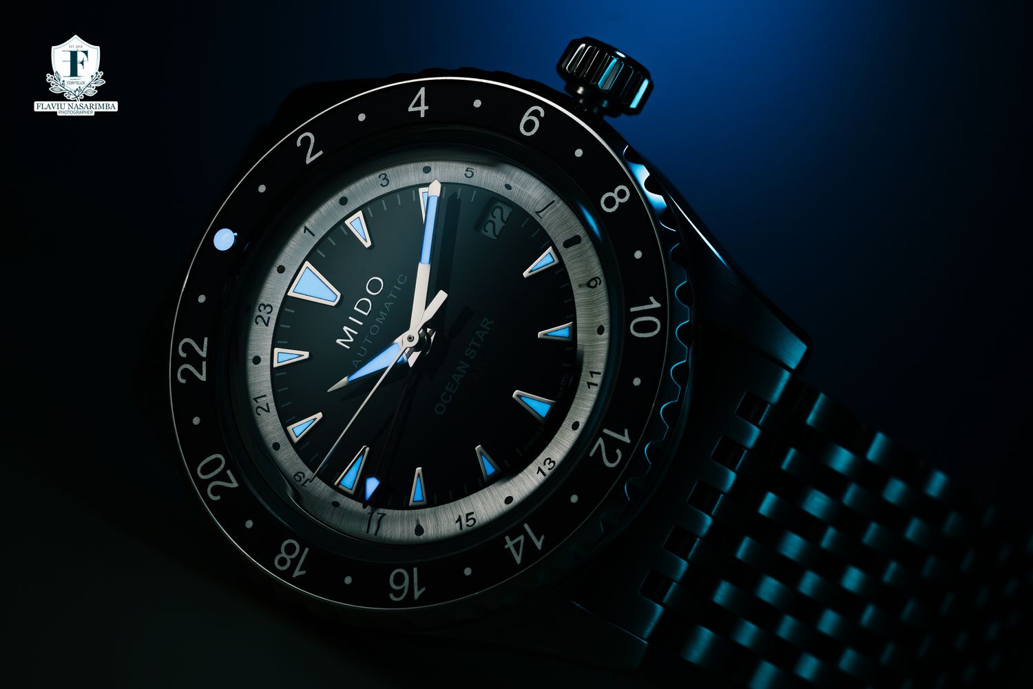Mido Ocean Star GMT watch with black dial and dual-time bezel on bracelet in blue dramatic lighting.