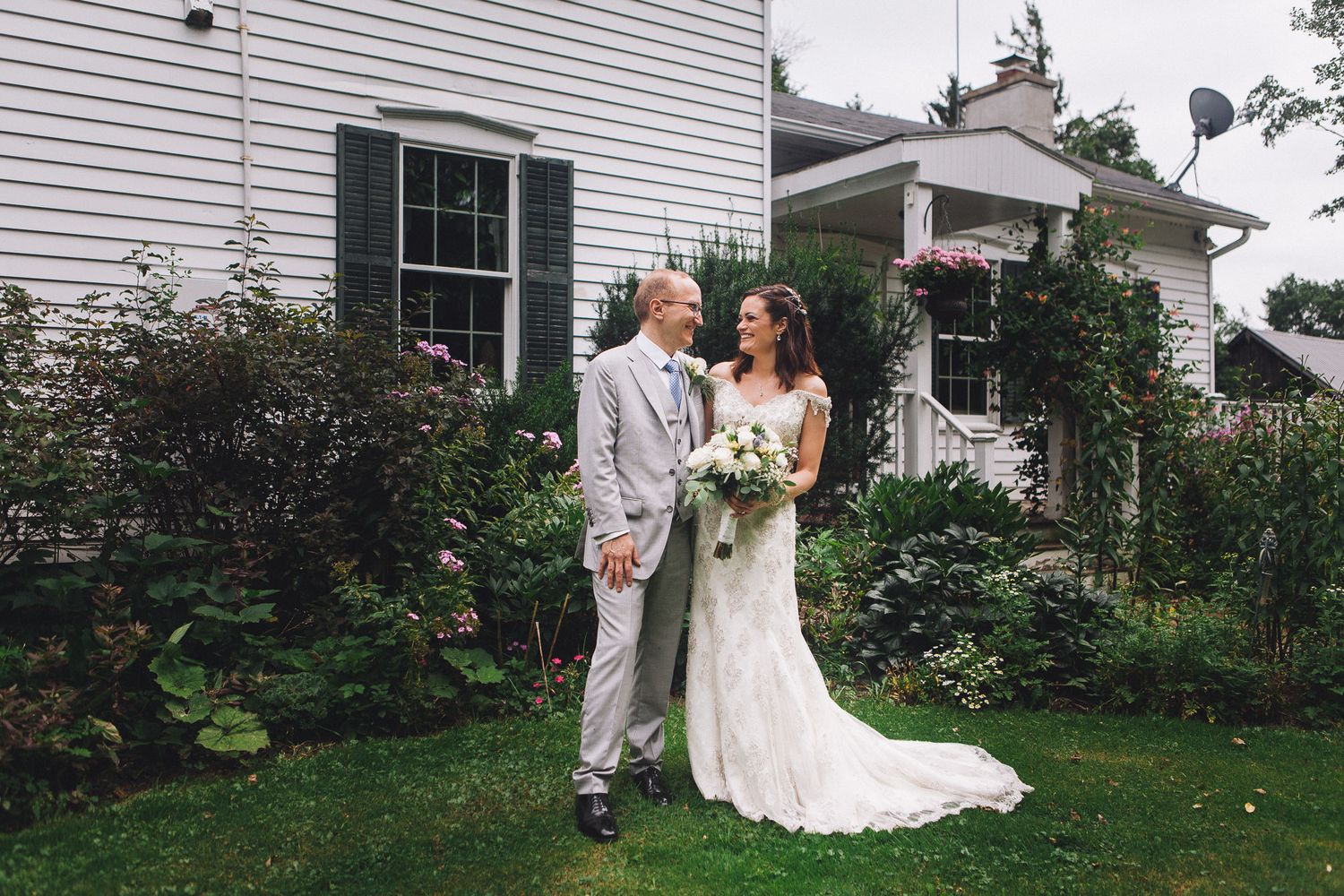 Bride and groom holding hands in lush garden beside white colonial house, bride in lace gown holding bouquet.