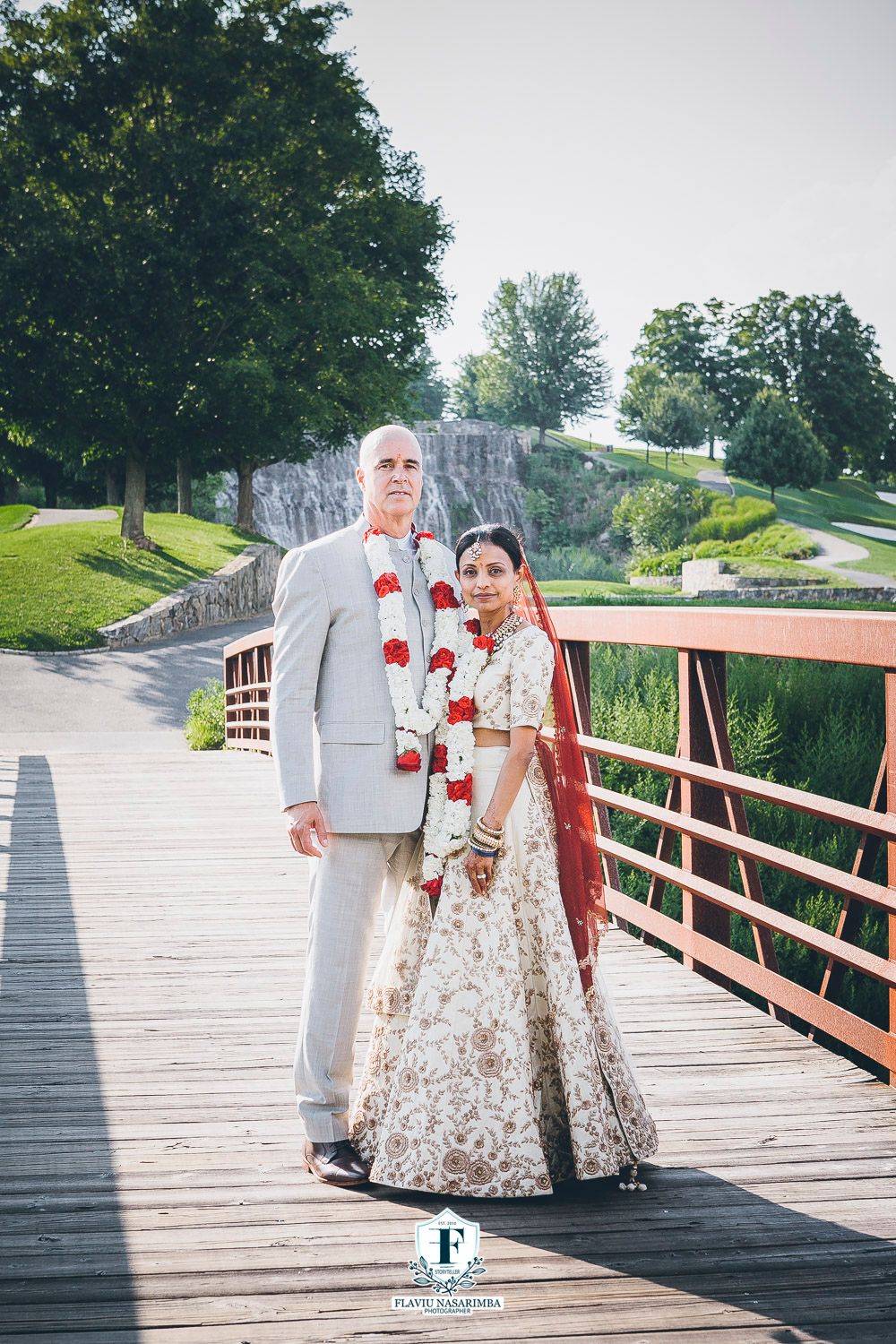 Couple in wedding attire posing on a bridge with a waterfall backdrop.