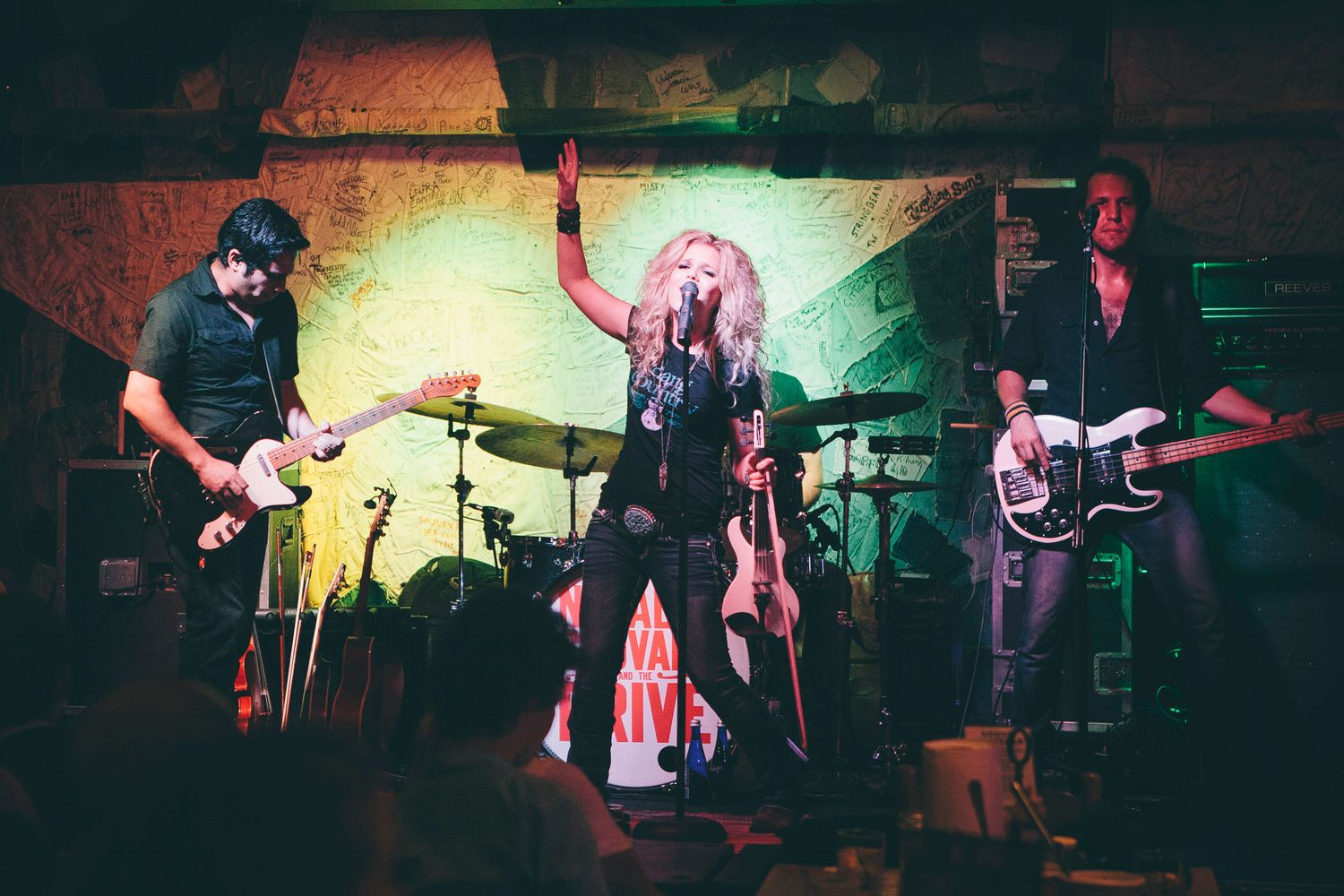 Female vocalist raises hand while performing on stage flanked by two electric guitarists.