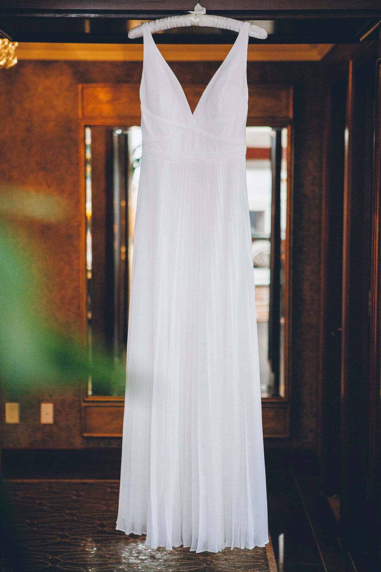 Elegant white pleated V-neck wedding gown hanging on a wooden door before the ceremony.