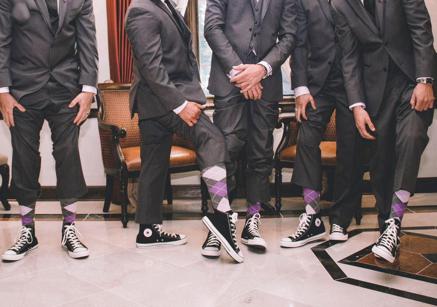 Groomsmen in gray suits wearing colorful purple argyle socks with black Converse sneakers at a wedding.