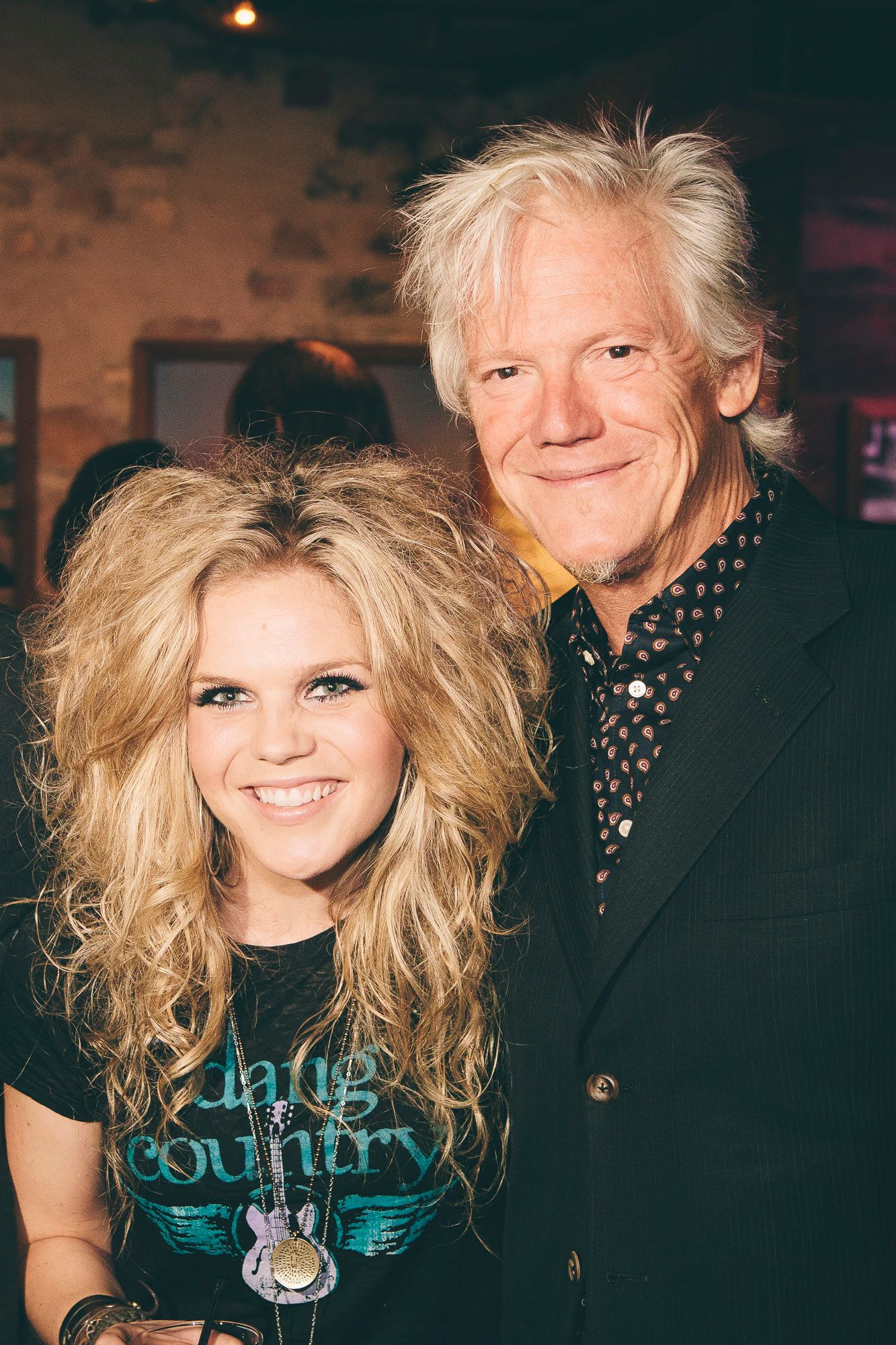 Young blonde woman and older man with silver hair smile together at a music event.