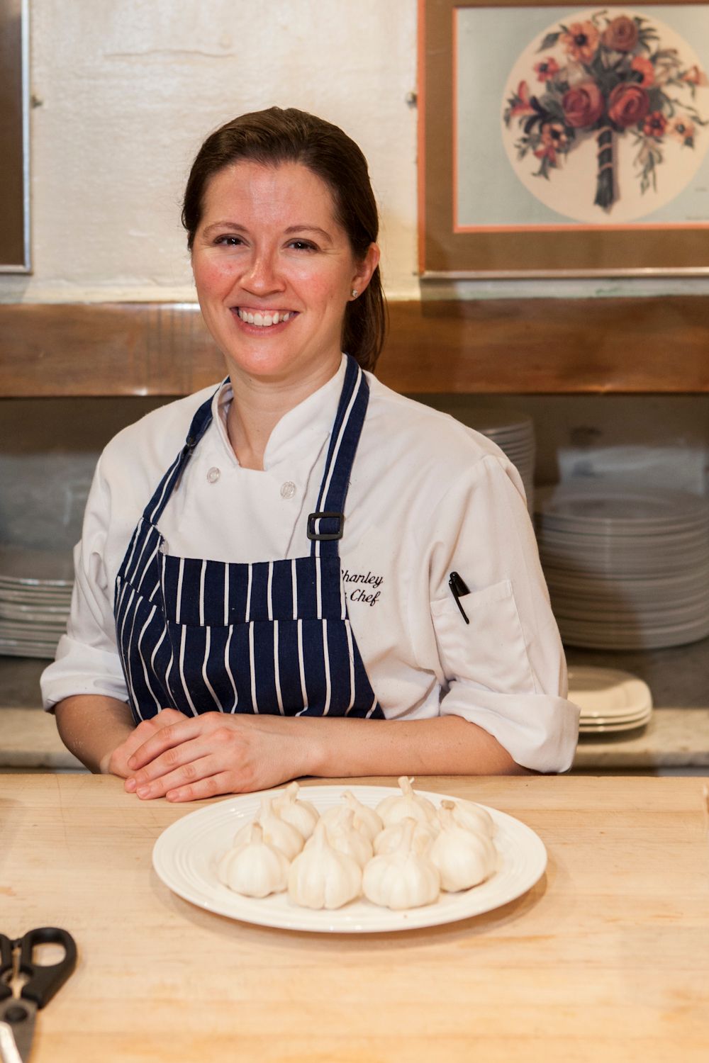 Smiling female chef in striped apron posing with a plate of white onions in a restaurant kitchen.