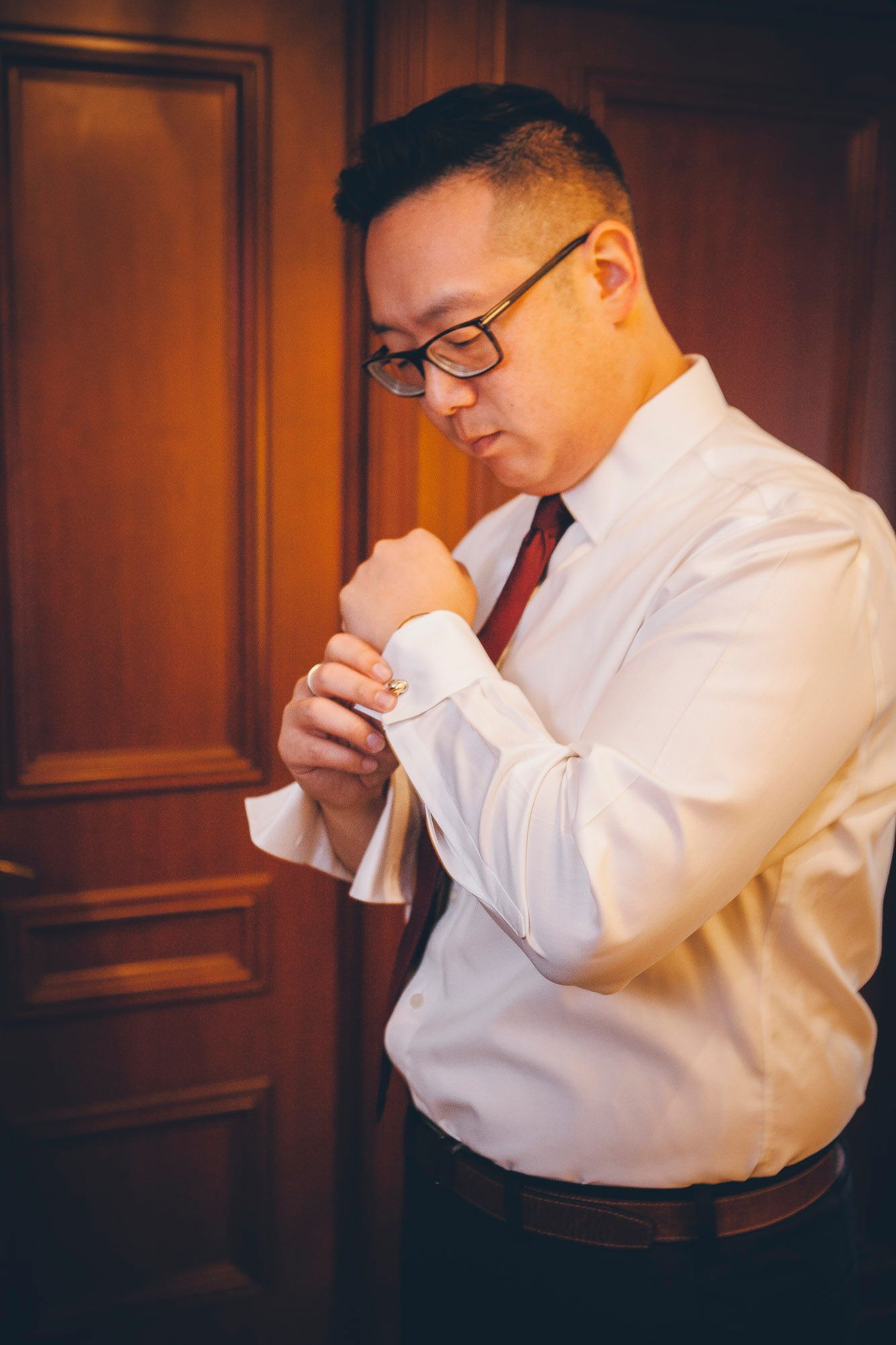 Groom in white shirt and red tie fastening his cufflinks before the wedding ceremony.
