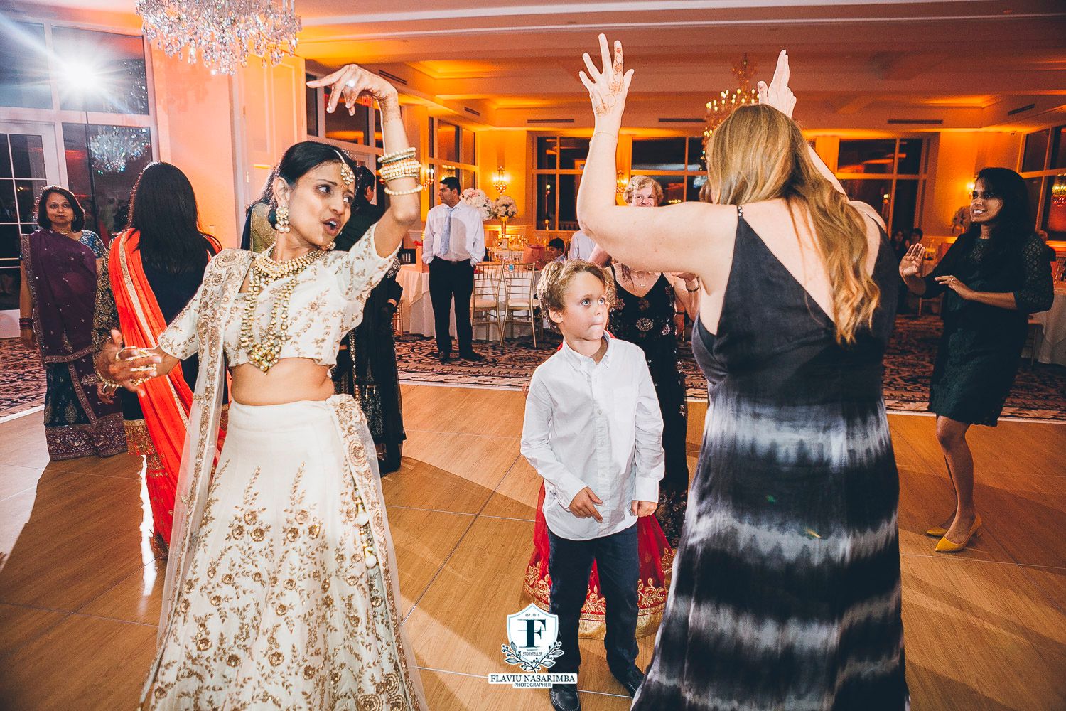 Indian bride in white lehenga dances with young child and guests at vibrant wedding reception celebration.