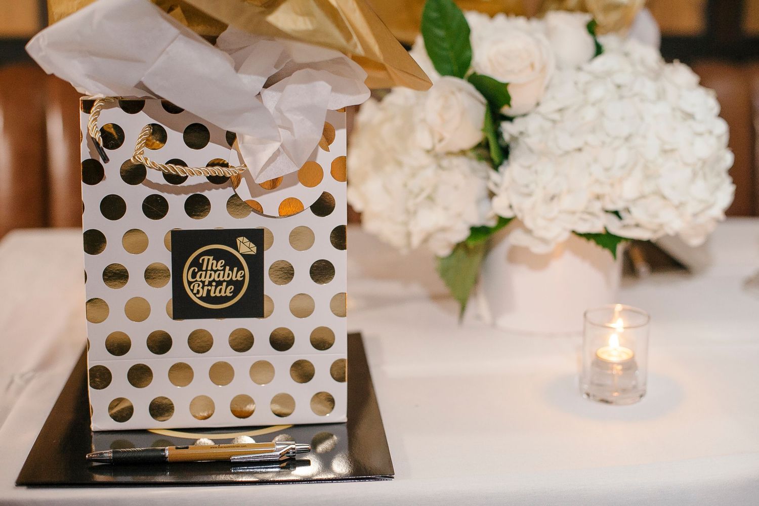 Gold polka dot gift bag beside white hydrangeas and candle at wedding reception.