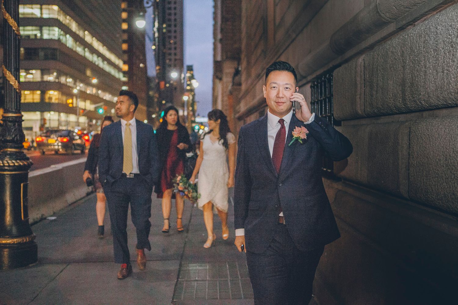 Groom leads wedding party along a city sidewalk at dusk in an urban setting.