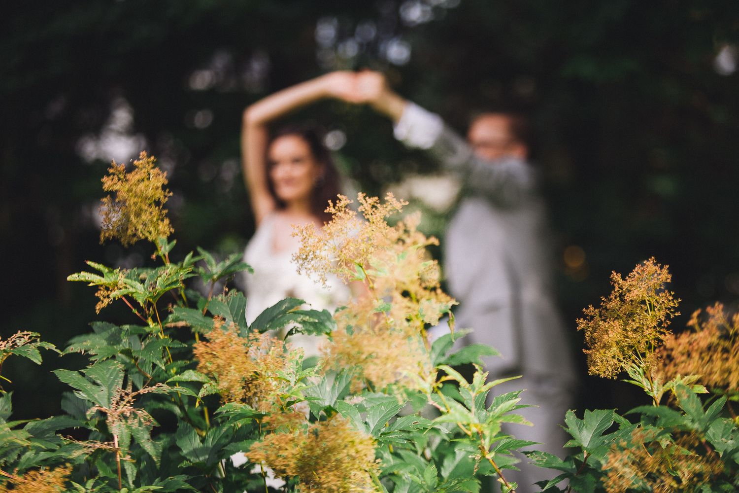 Bride and groom forming a heart with their arms behind golden wildflowers in soft garden light.