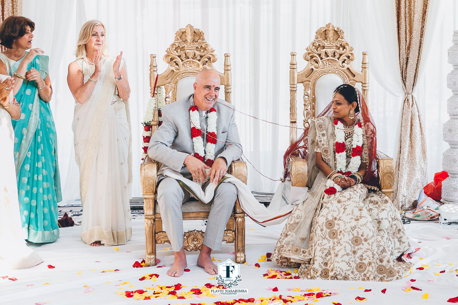 Interracial couple in traditional Indian wedding attire seated on ornate gold thrones during Hindu ceremony.