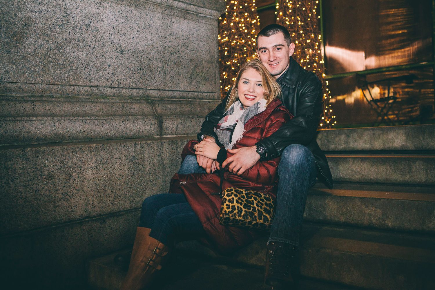Happy couple sits closely together on stone steps beside glowing Christmas tree at night.