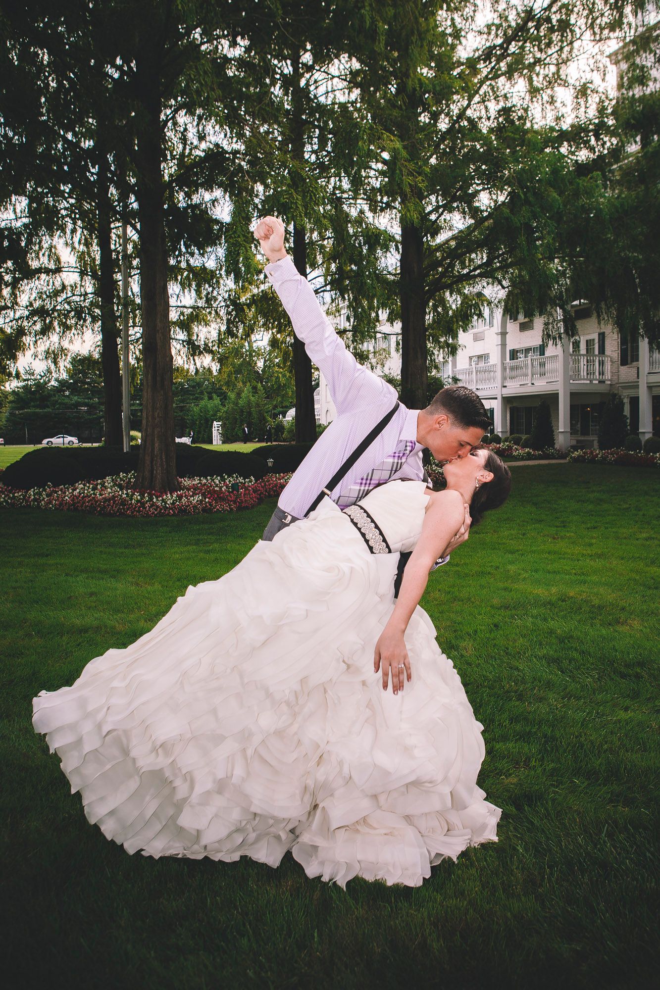 Groom dipping bride for a romantic kiss on green lawn, bride in ruffled white gown outdoors.