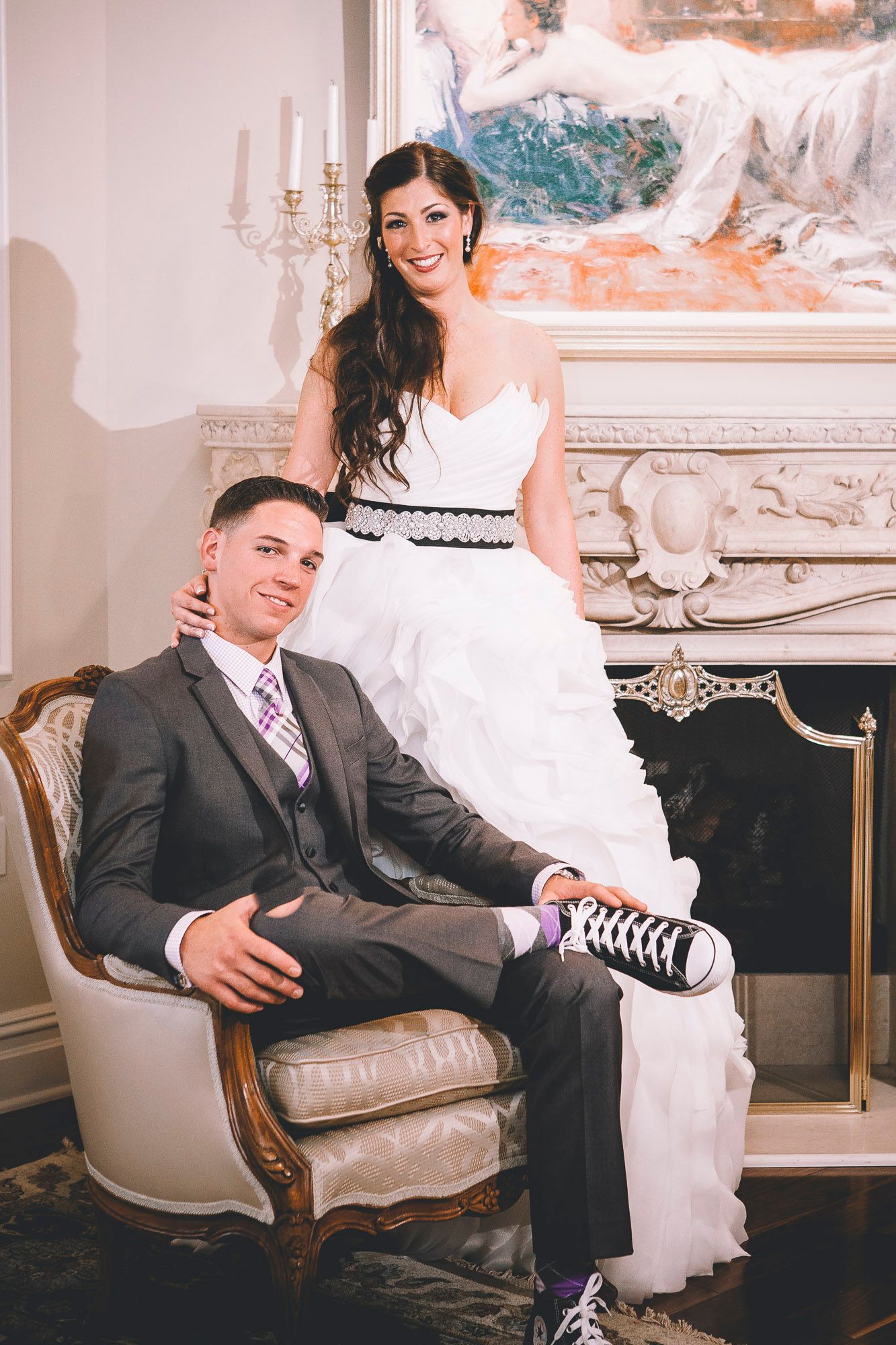 Bride and groom pose by fireplace, groom seated in armchair, bride wearing black Converse sneakers.