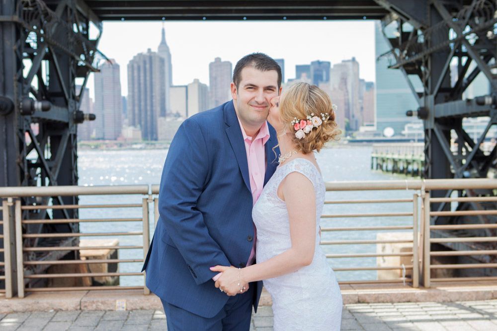 Groom in navy suit and bride in white lace dress smile at each other near industrial bridge structure.