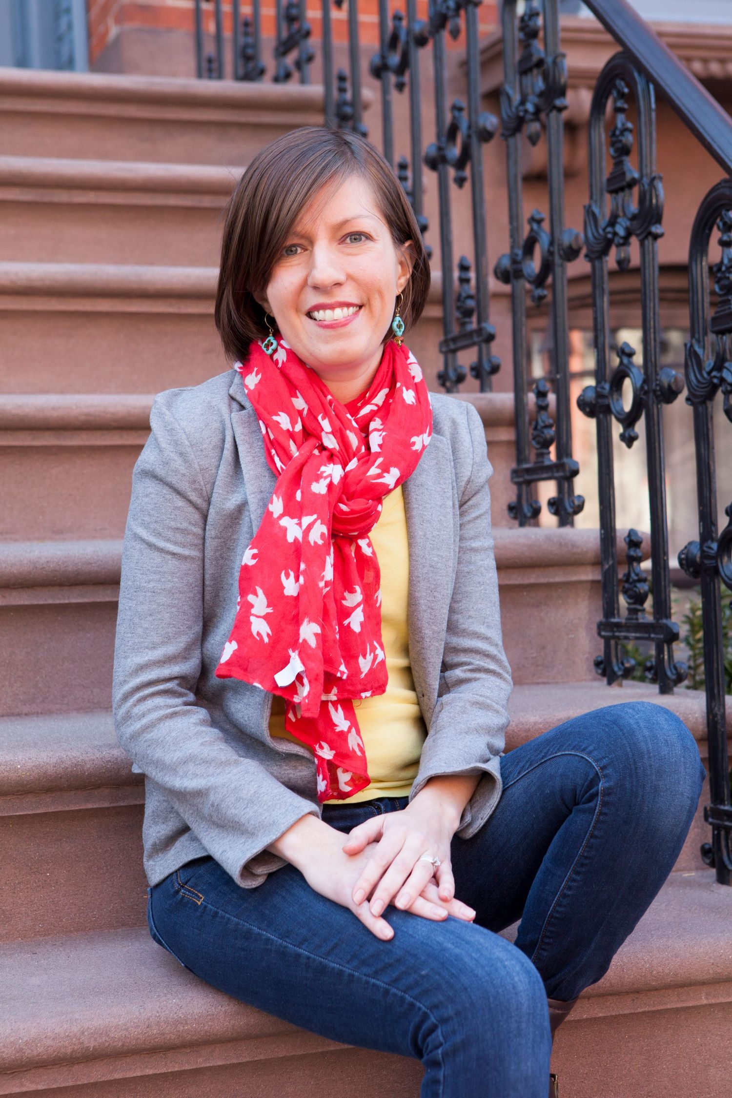 Smiling woman in gray blazer, red scarf, and jeans sitting on brownstone steps.