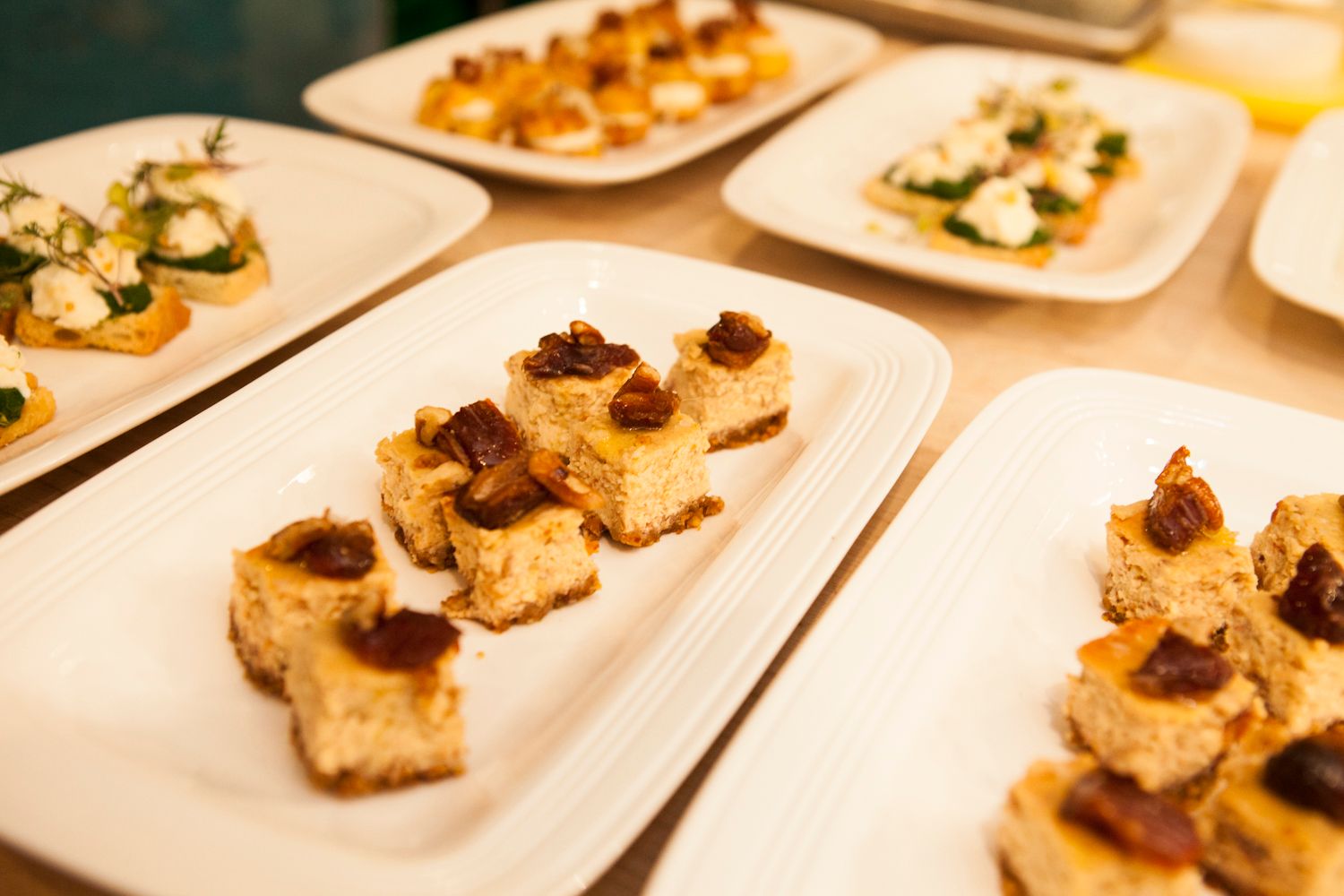 Rows of white square plates with toasted bread cubes topped with dried fruit at a catered event.