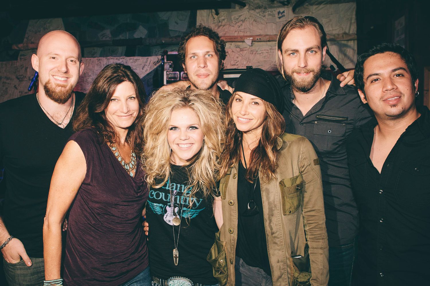 Group of seven musicians smiling together for a photo backstage at a live music venue.