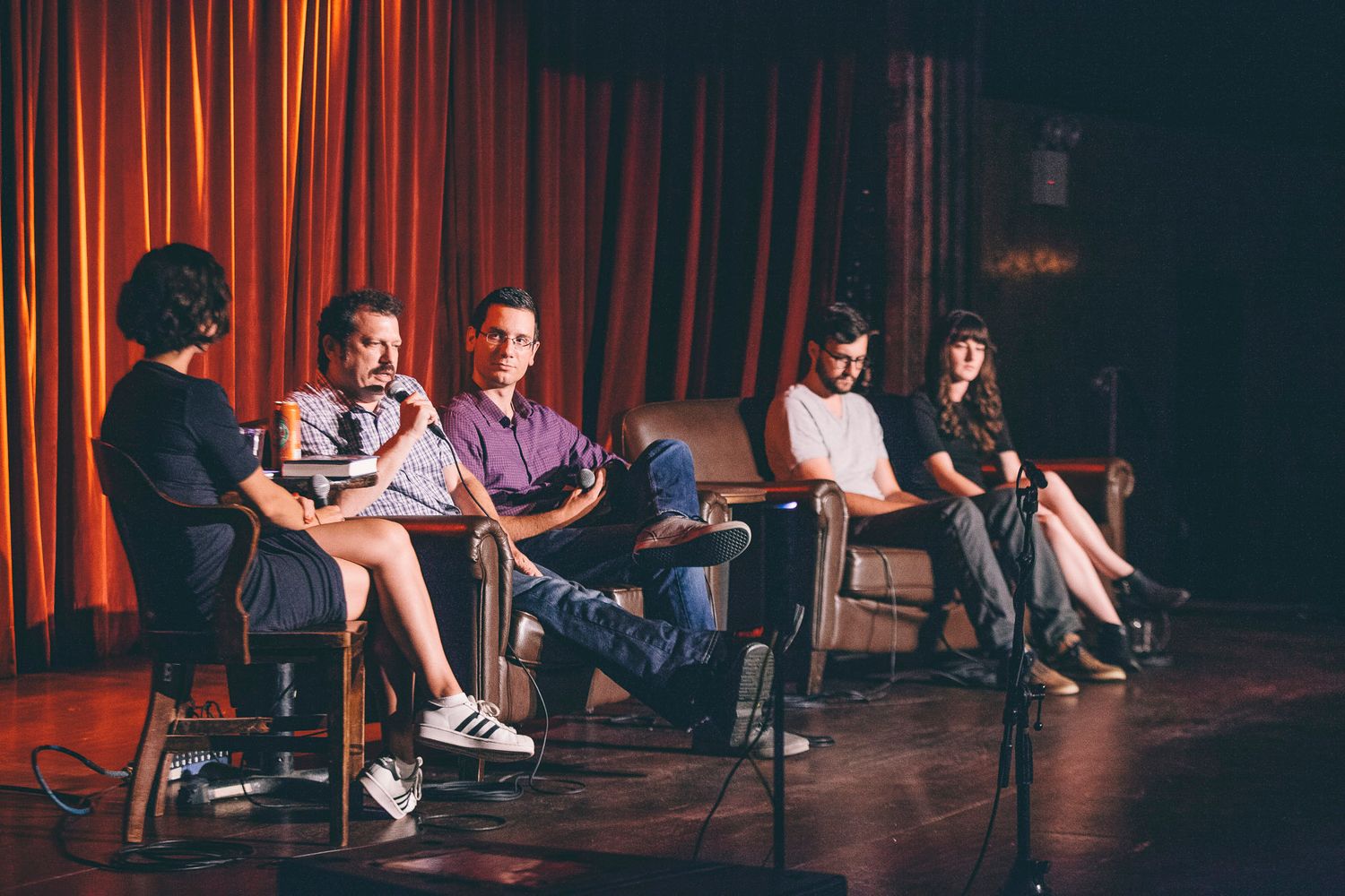 Panel discussion with five people seated on stage in front of red curtains at a comedy venue.