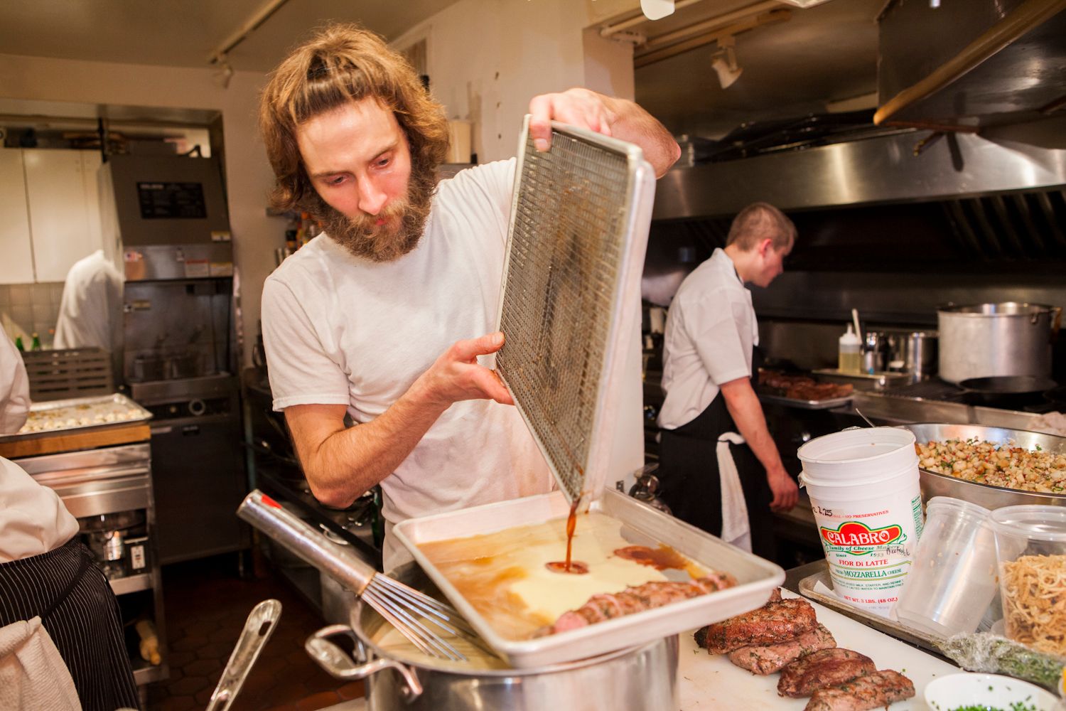 Bearded male chef straining sauce over a pan of food in a busy professional kitchen.