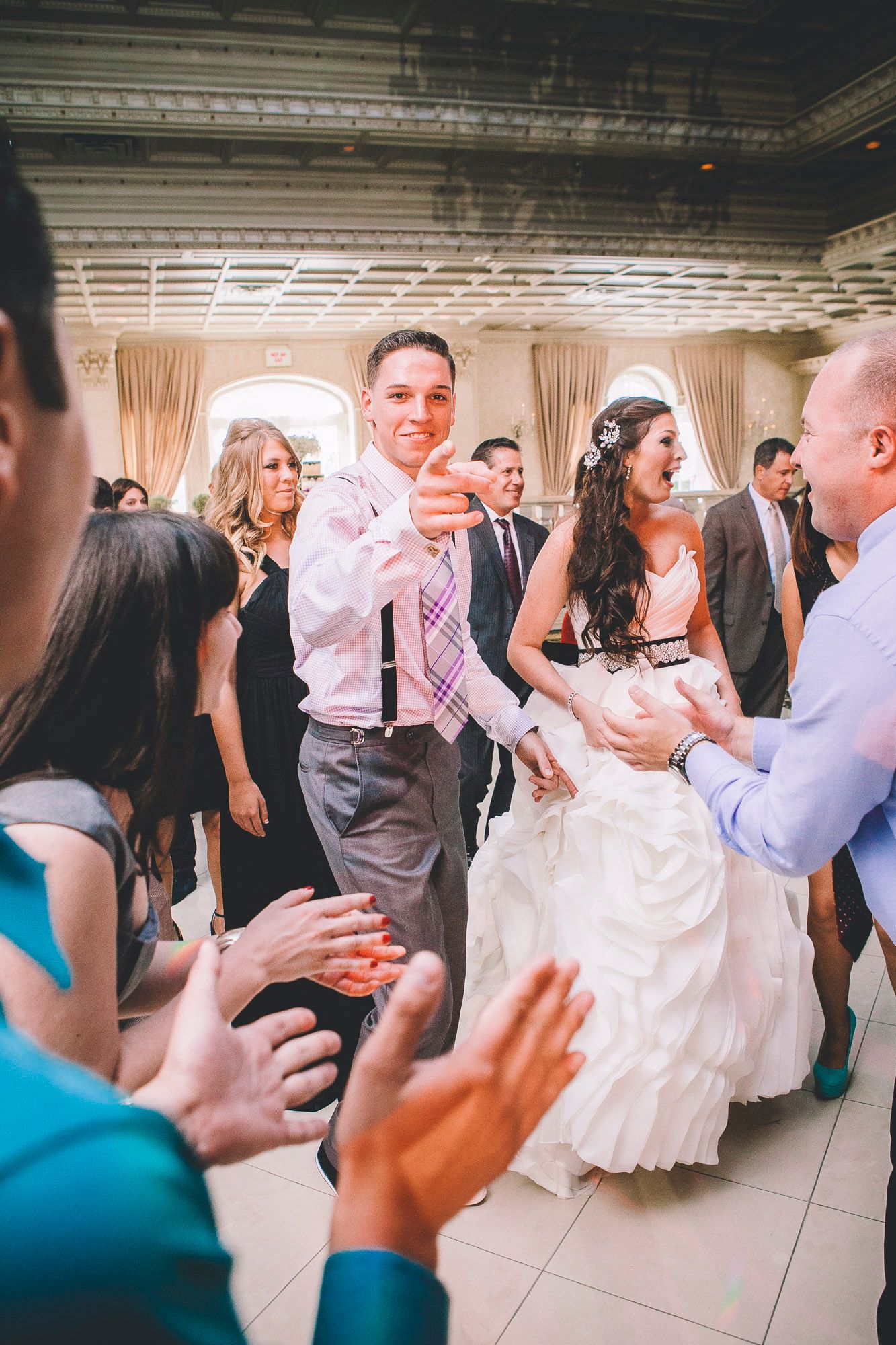 Bride and groom dancing joyfully with wedding guests clapping on the dance floor at their reception.
