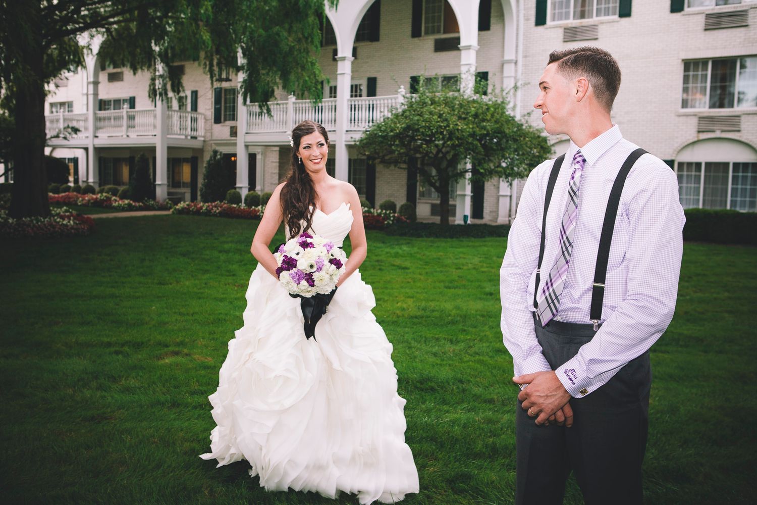 Bride holding purple bouquet faces groom in first look wedding moment on green lawn.