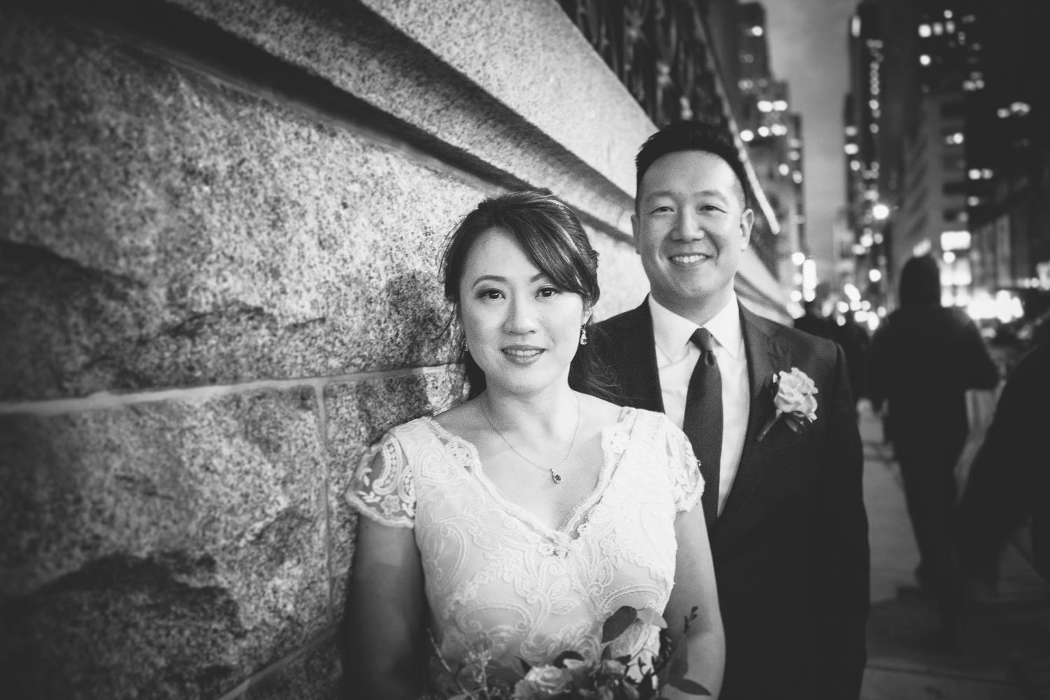 Newlyweds pose against a stone wall on a city street in an elegant black and white portrait.