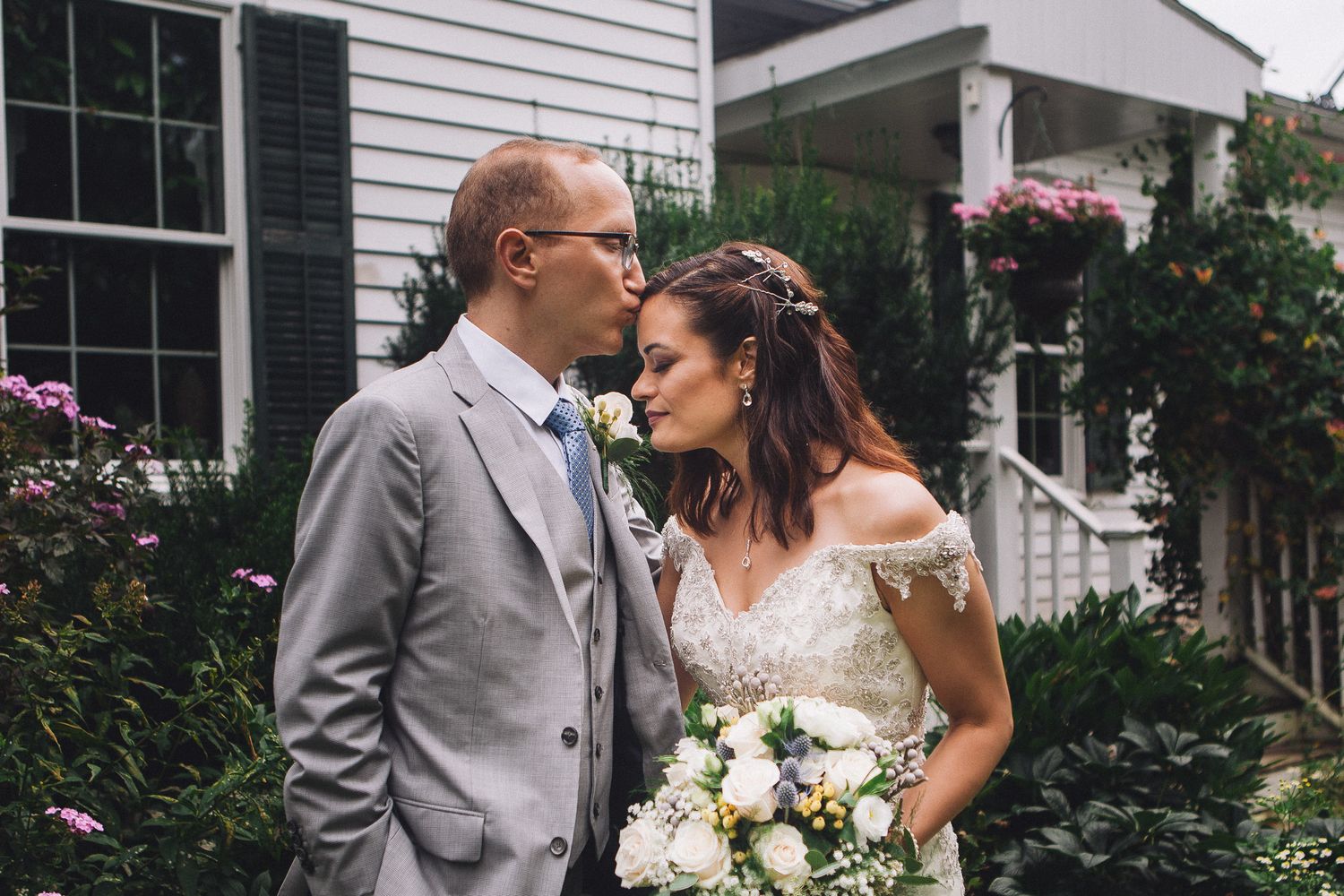 Bride and groom sharing a romantic moment near a white house surrounded by lush garden flowers.