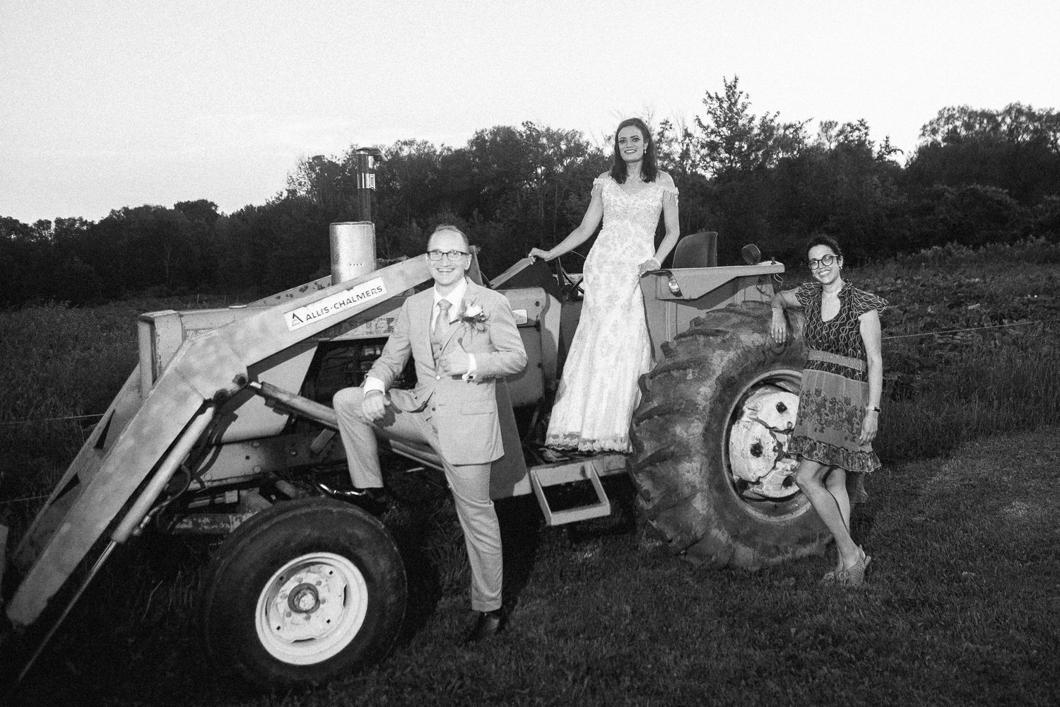 Black and white photo of bride, groom, and guest posing on a vintage farm tractor at sunset.