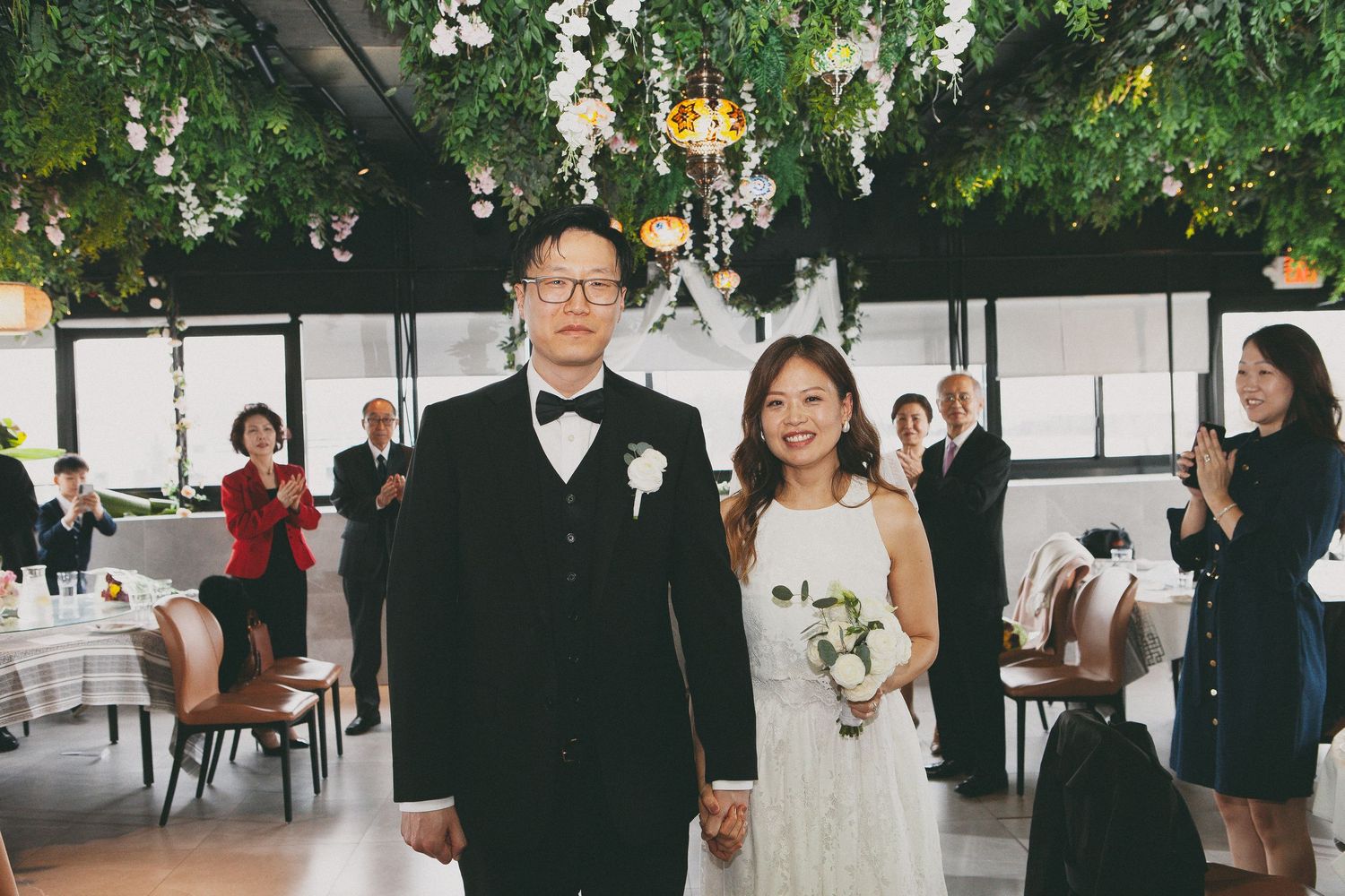 Newlywed Asian couple walks hand in hand through restaurant venue as guests celebrate with applause.
