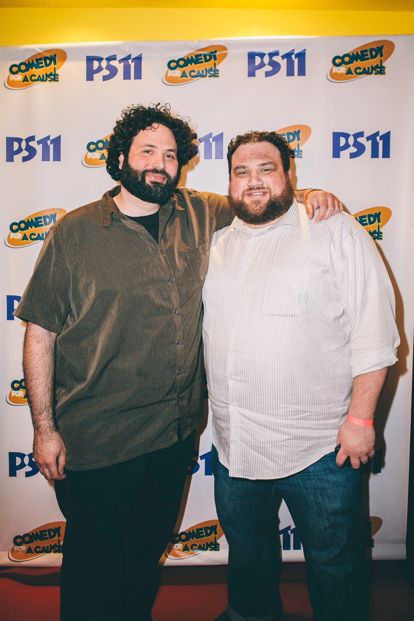 Two men smiling together at PS11 Comedy for a Cause charity fundraiser event backdrop.