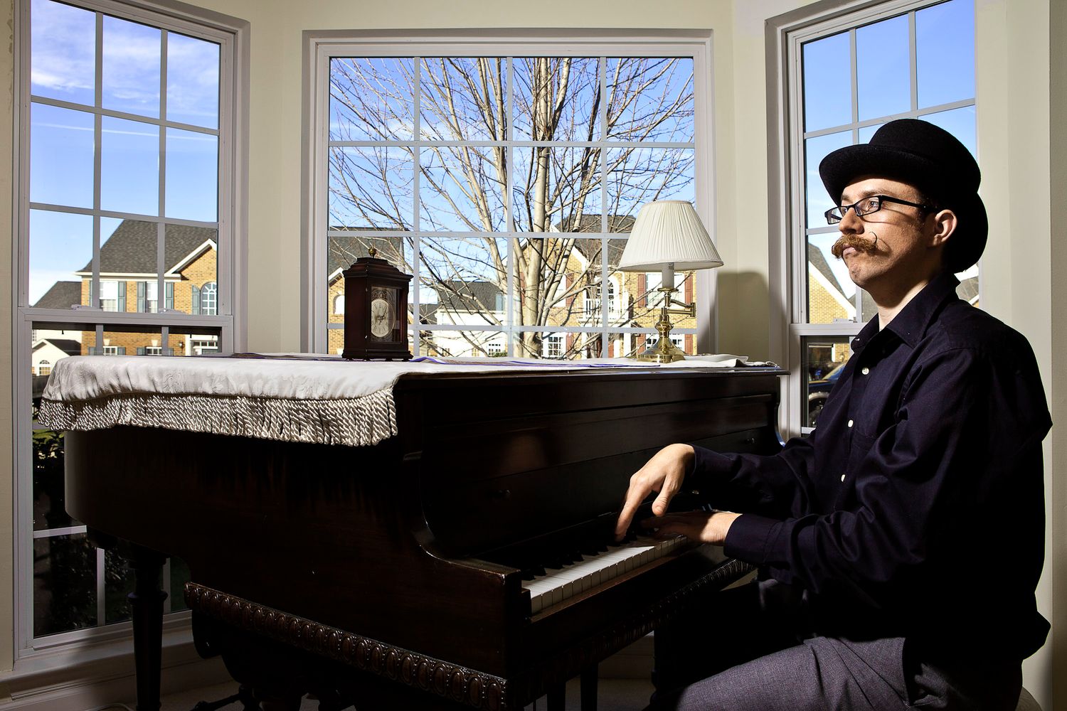 Young man in black hat sitting at a grand piano by large windows in a bright living room.