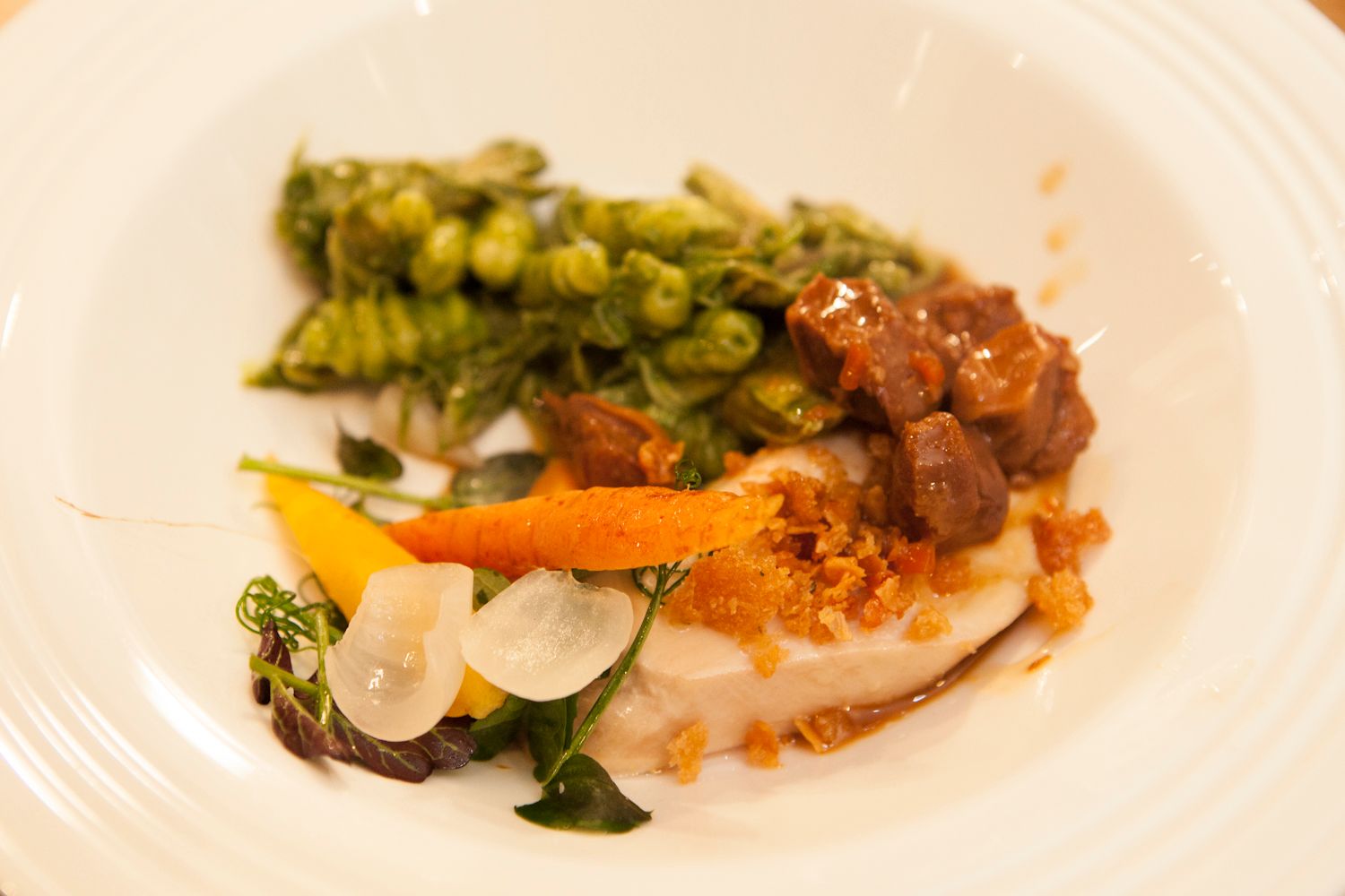 Elegant plated dish with white fish, green pea gnocchi, braised meat, roasted carrots, and fresh herbs on a white plate.