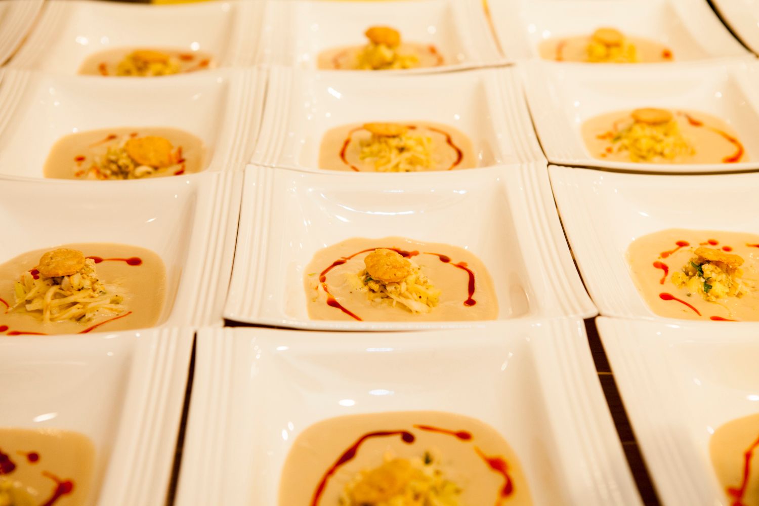 Rows of white square dishes with creamy soup garnished with red sauce and herbs at a catered event.