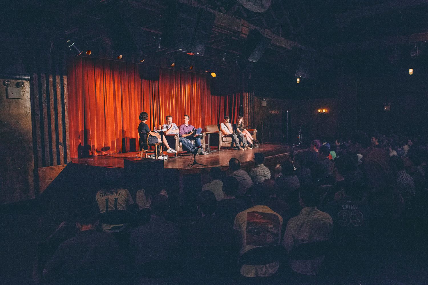Panel discussion on stage with red curtain backdrop in dimly lit theater venue.