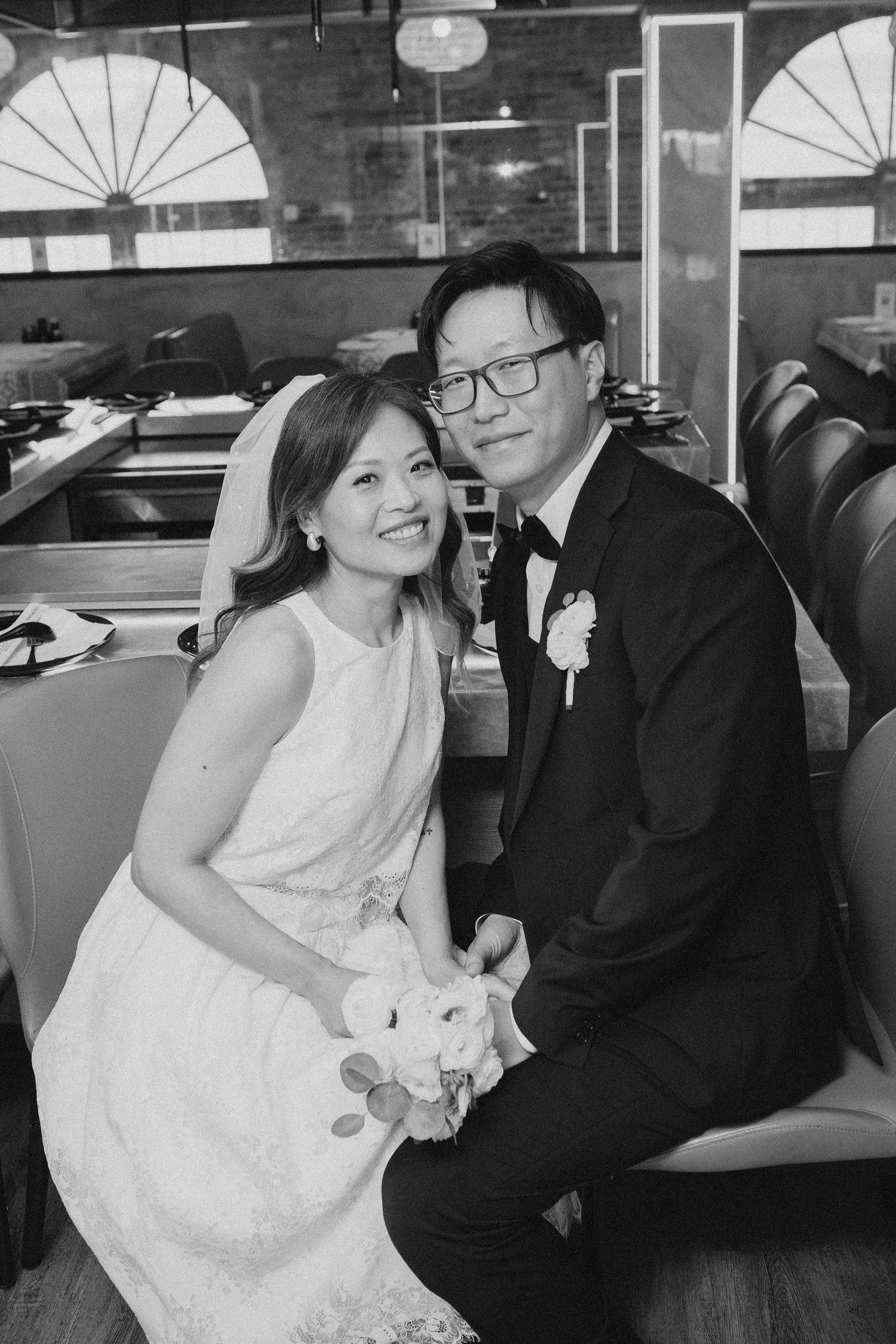Black and white portrait of smiling Asian bride and groom seated together holding bridal bouquet.