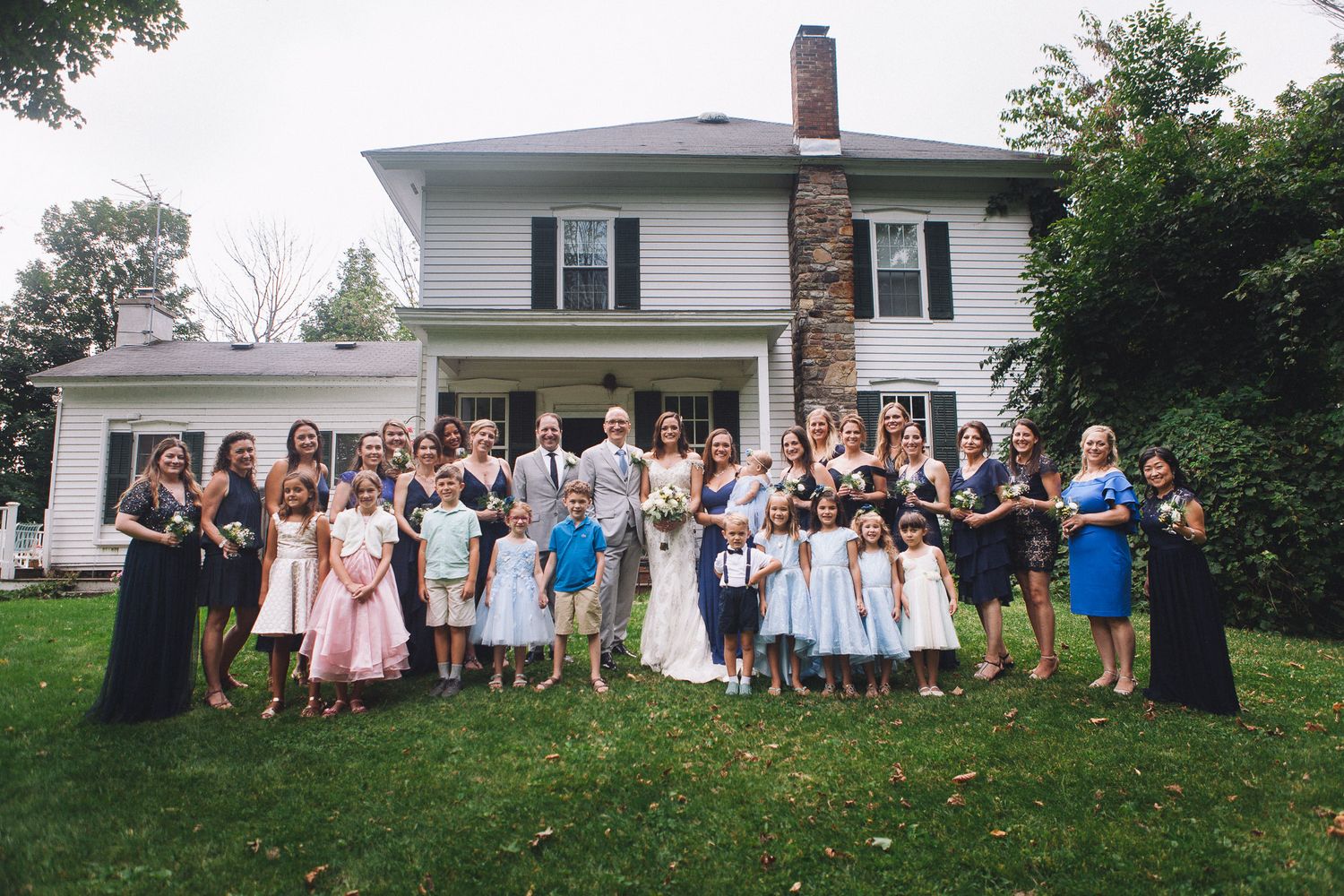 Large wedding party posing together in front of a white two-story house on a cloudy day.