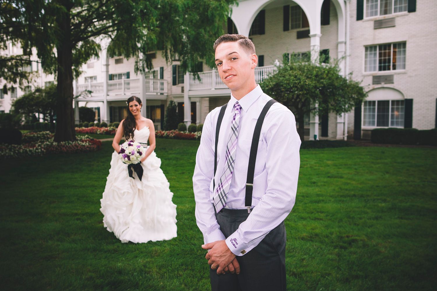 Groom in suspenders stands in foreground as bride approaches in ruffled gown outdoors.