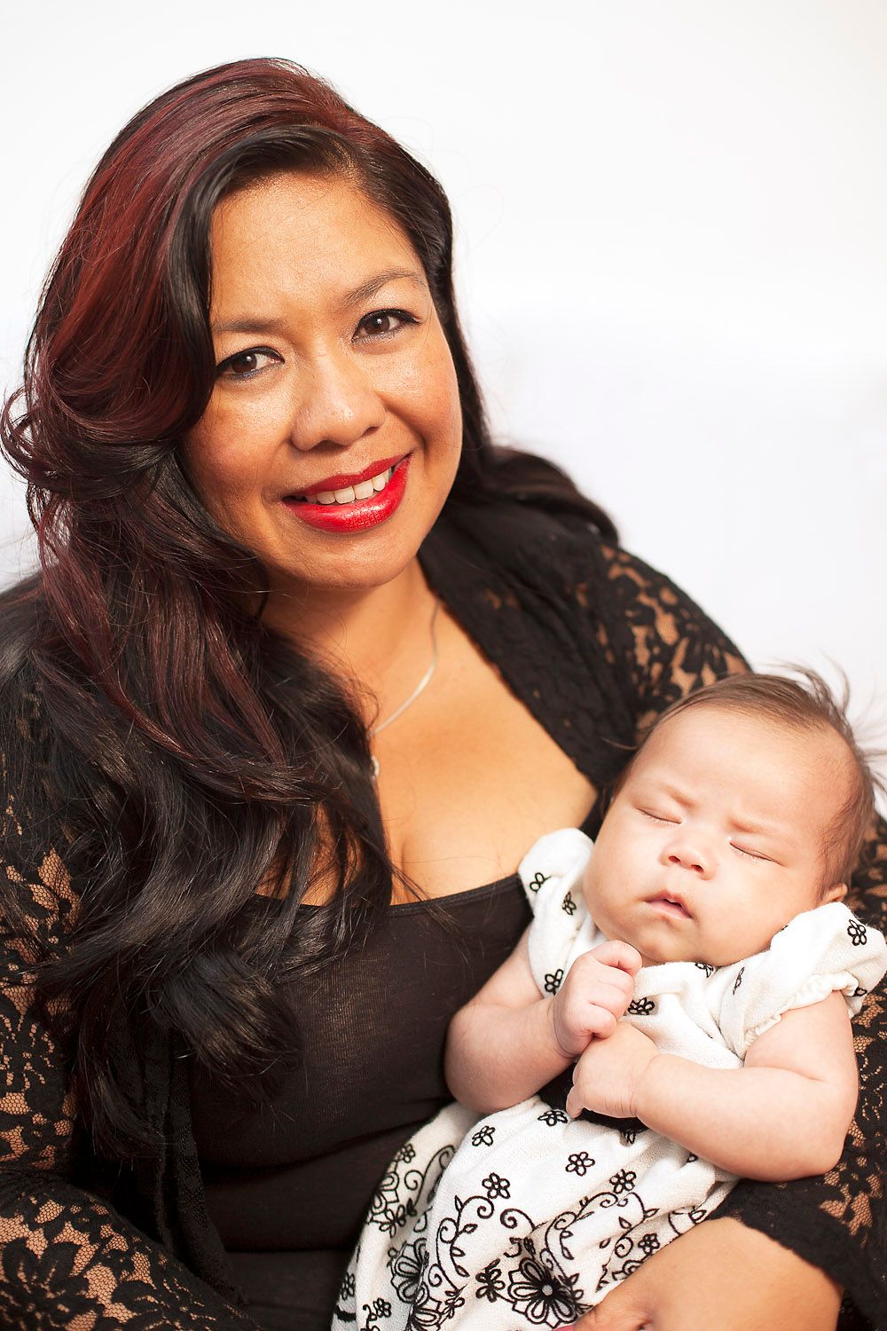 Smiling Asian woman with red highlights holding sleeping newborn baby in studio portrait.