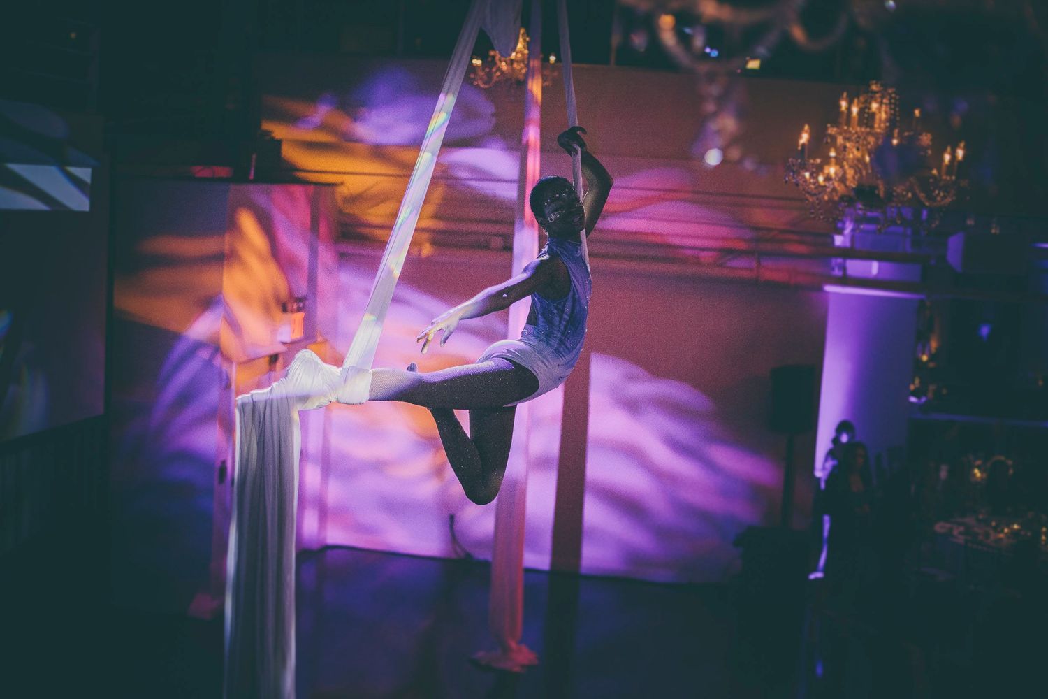 An aerial acrobat performs a seated split pose on white silk fabric under warm chandelier lighting.