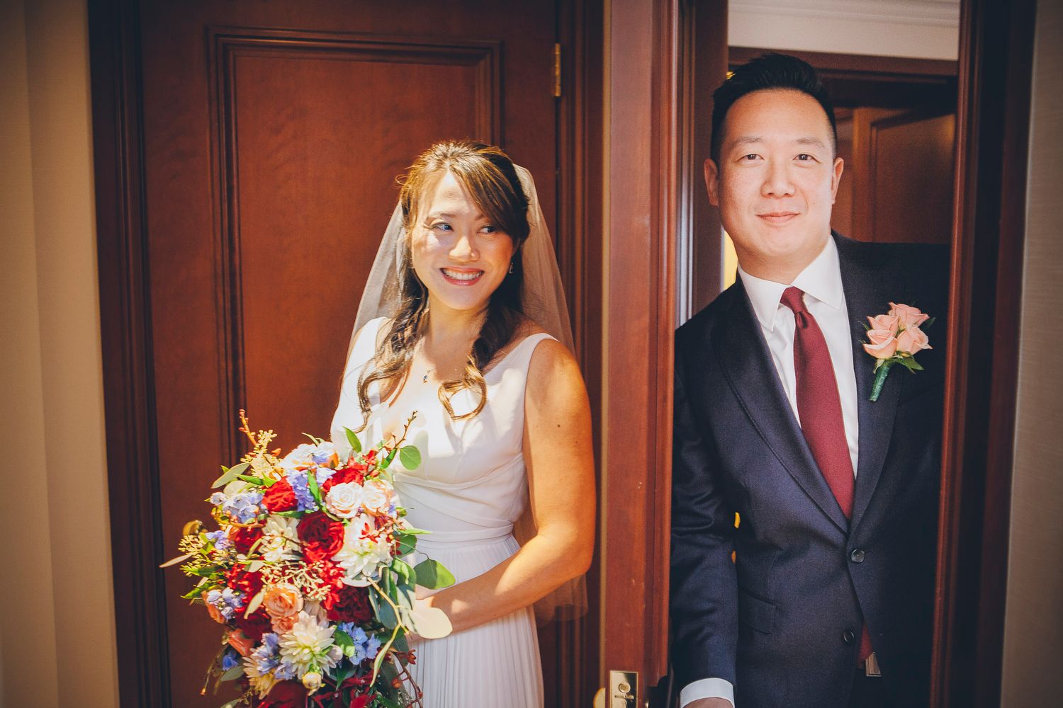 Bride and groom smiling together in a doorway, bride holding a vibrant cascading floral bouquet.